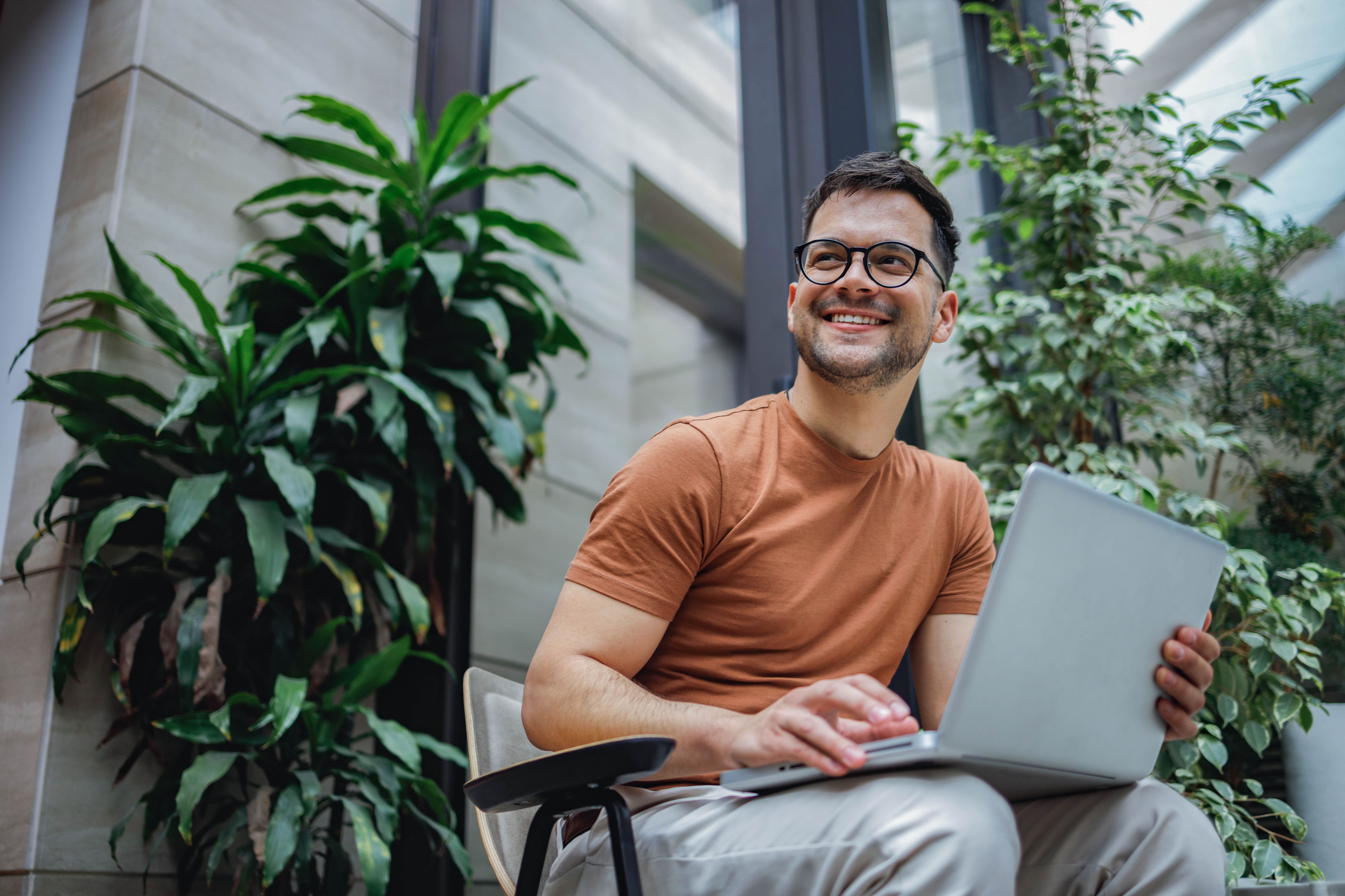 Mann mit Brille sitzt in einem modernen Raum, lächelt und arbeitet an einem Laptop, umgeben von Pflanzen.