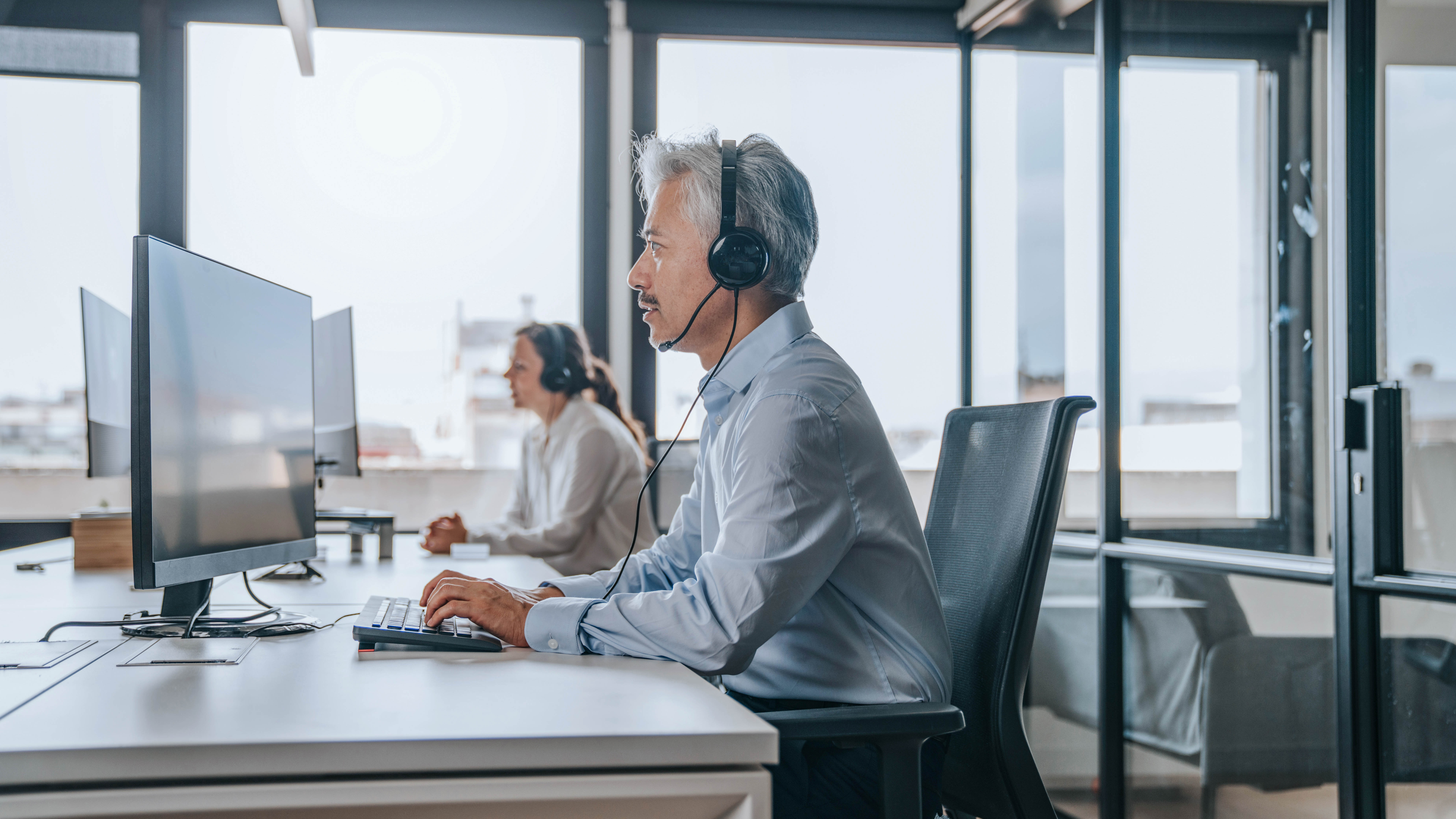 Mann mit Headset sitzt an einem Schreibtisch vor einem Computer, im Hintergrund eine Frau am Arbeitsplatz.
