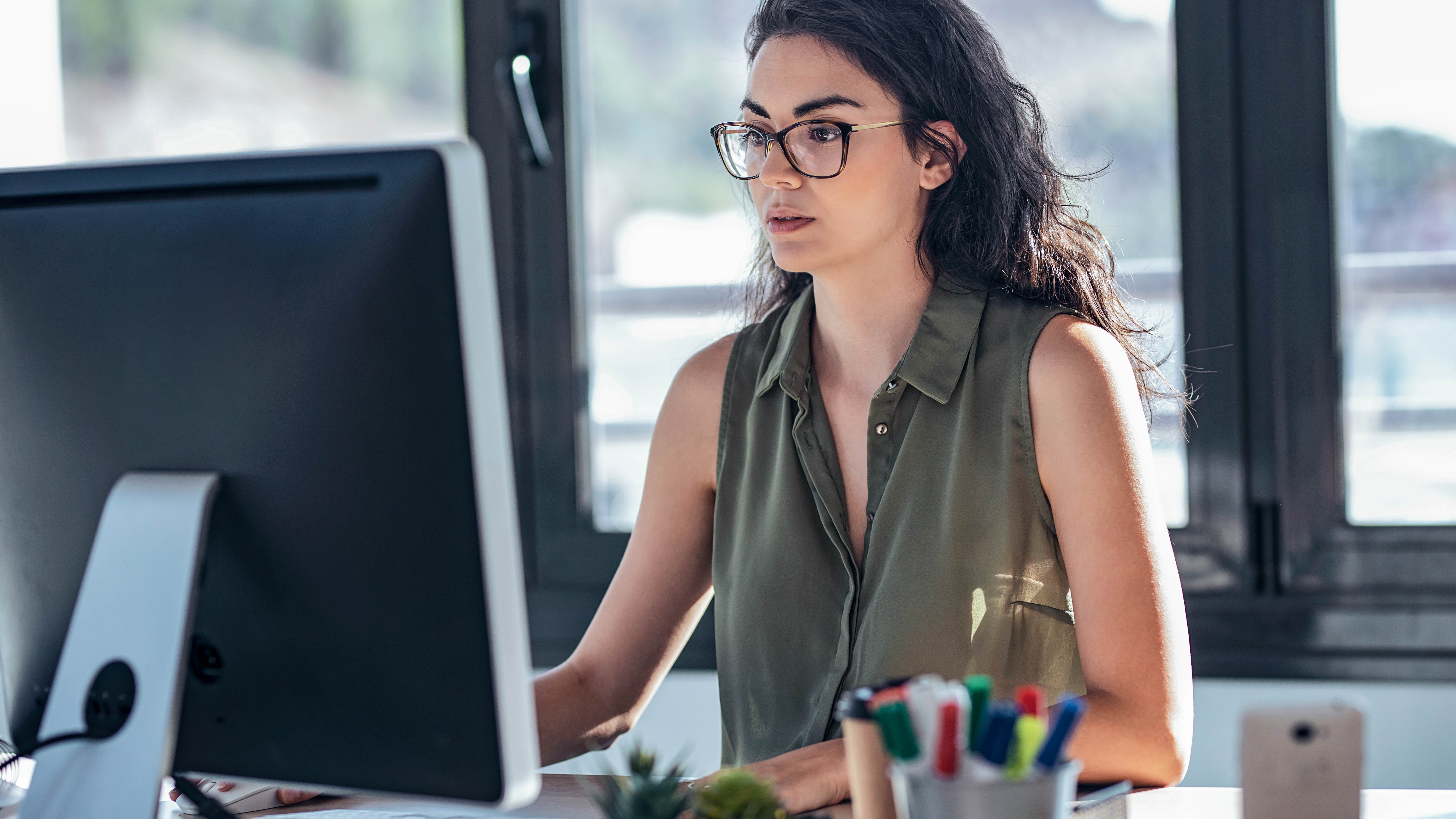 Frau mit Brille sitzt an einem Schreibtisch und arbeitet konzentriert am Computer.
