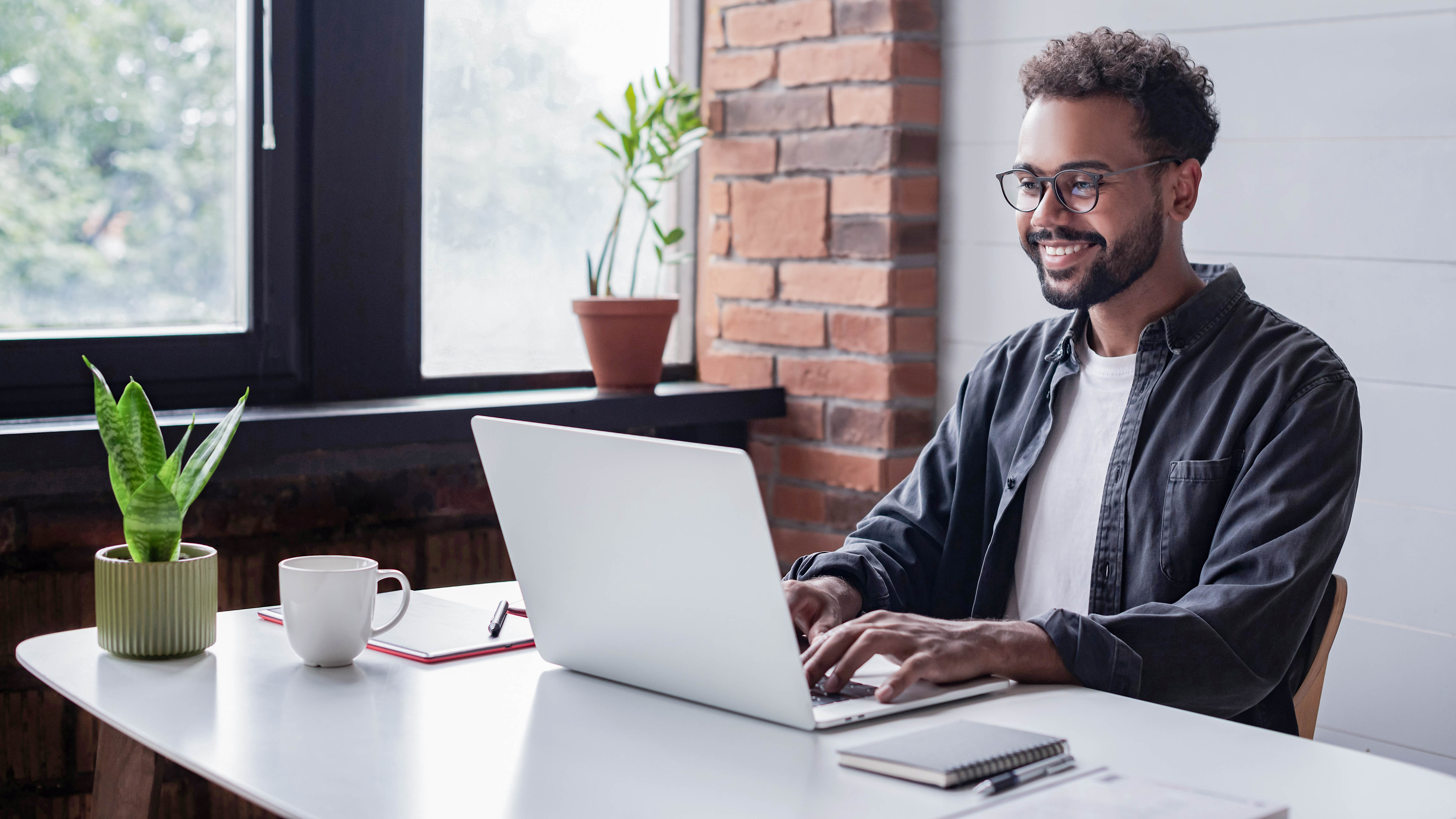 Mann mit Brille sitzt an einem Tisch und arbeitet an einem Laptop, neben ihm eine Tasse und eine Pflanze.