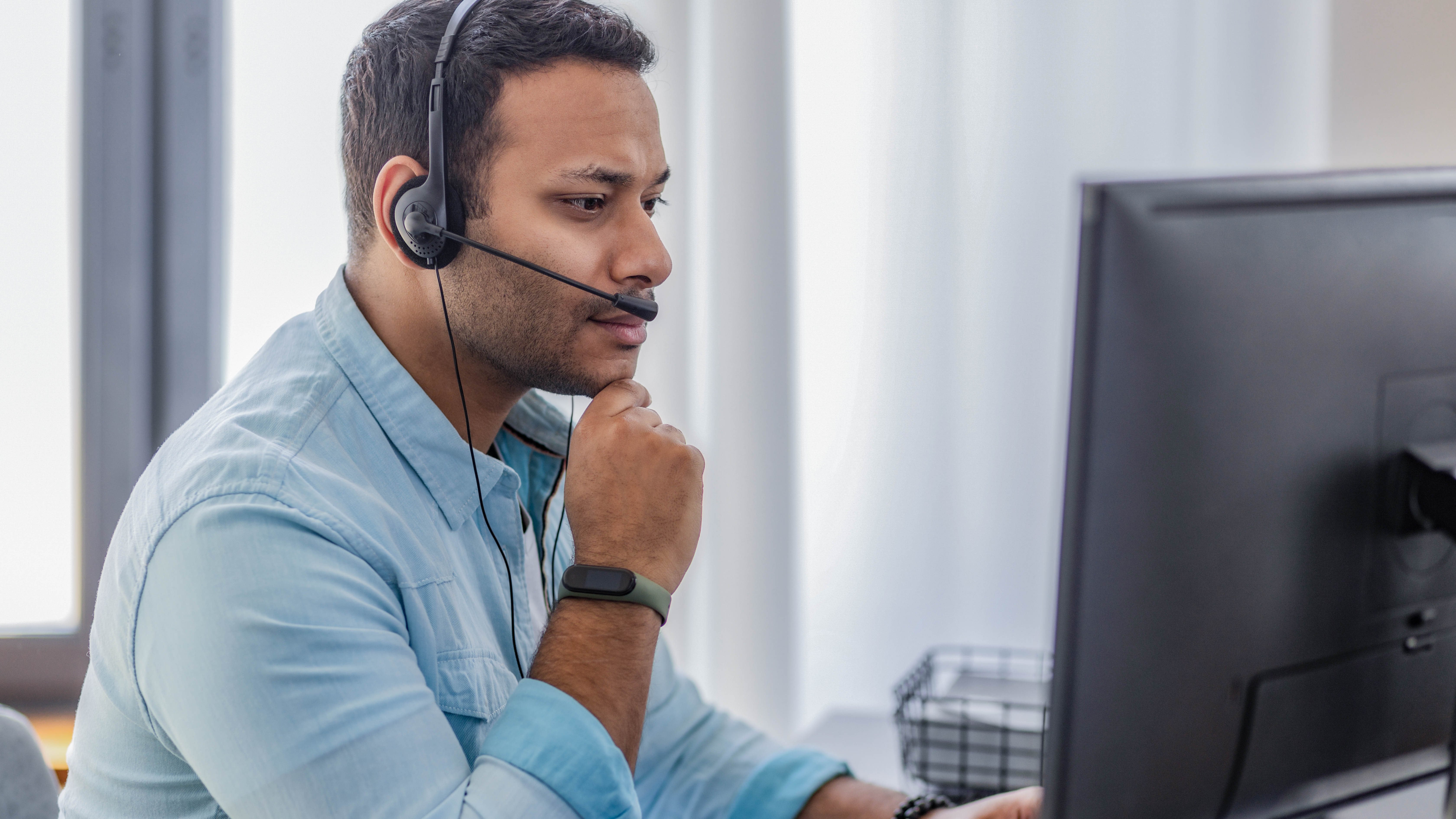 Mann mit Headset sitzt vor einem Computer und schaut nachdenklich auf den Bildschirm.