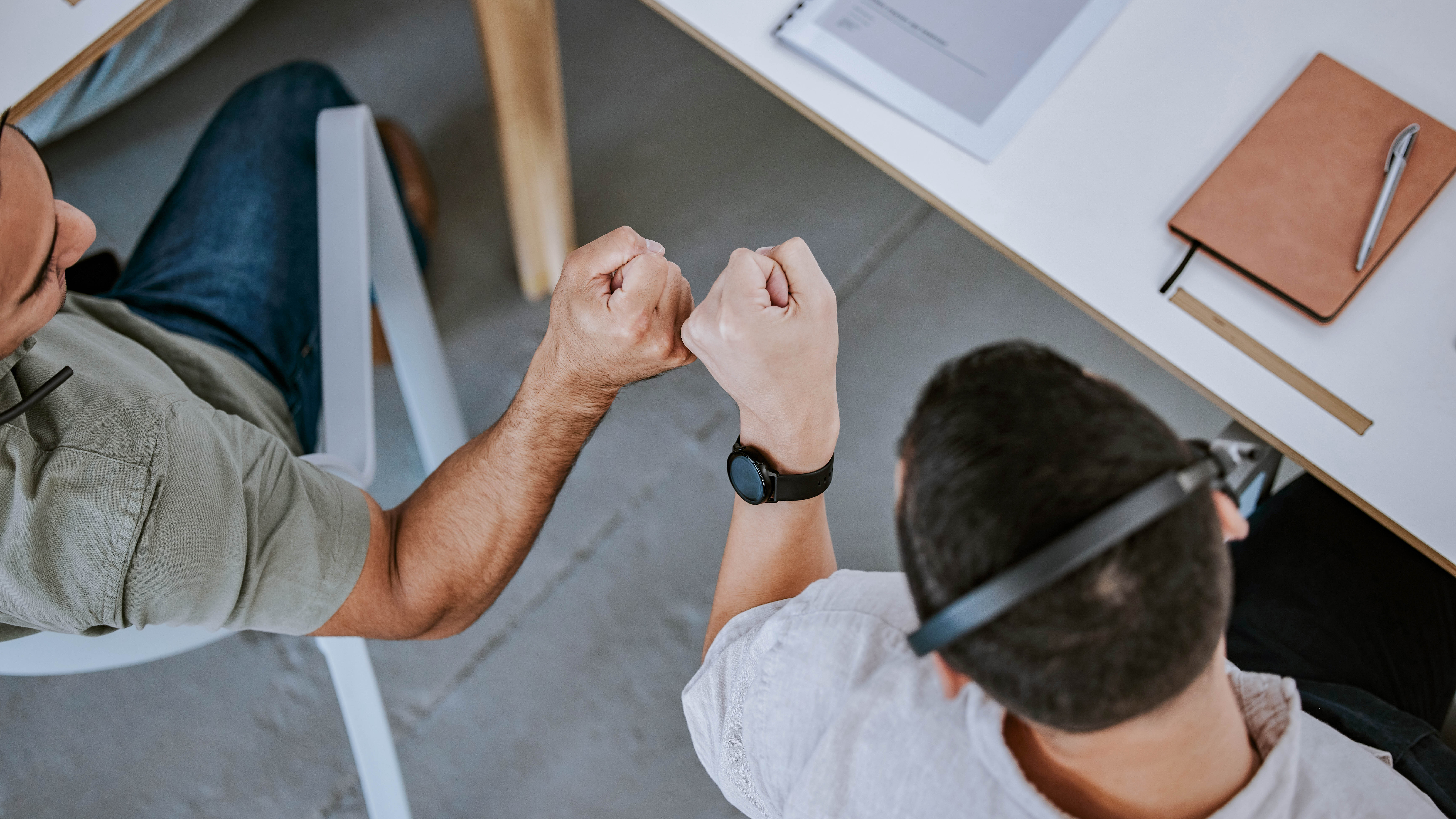 Zwei Personen geben sich fist bumps über einem Tisch, auf dem Schreibutensilien und Unterlagen liegen.