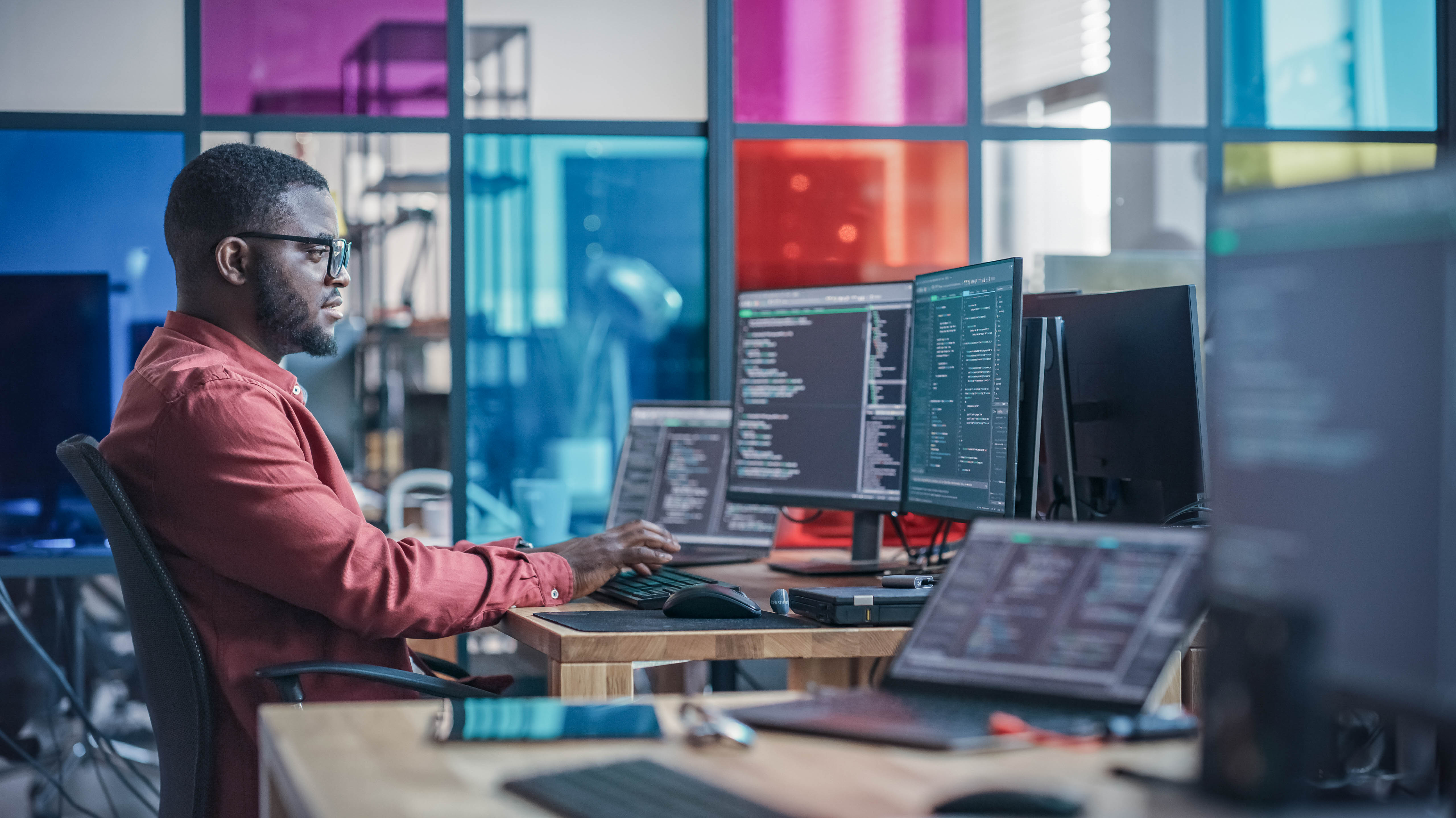 Ein Mann sitzt an einem Schreibtisch mit mehreren Monitoren, die Programmiercode anzeigen, in einem modernen Büro.
