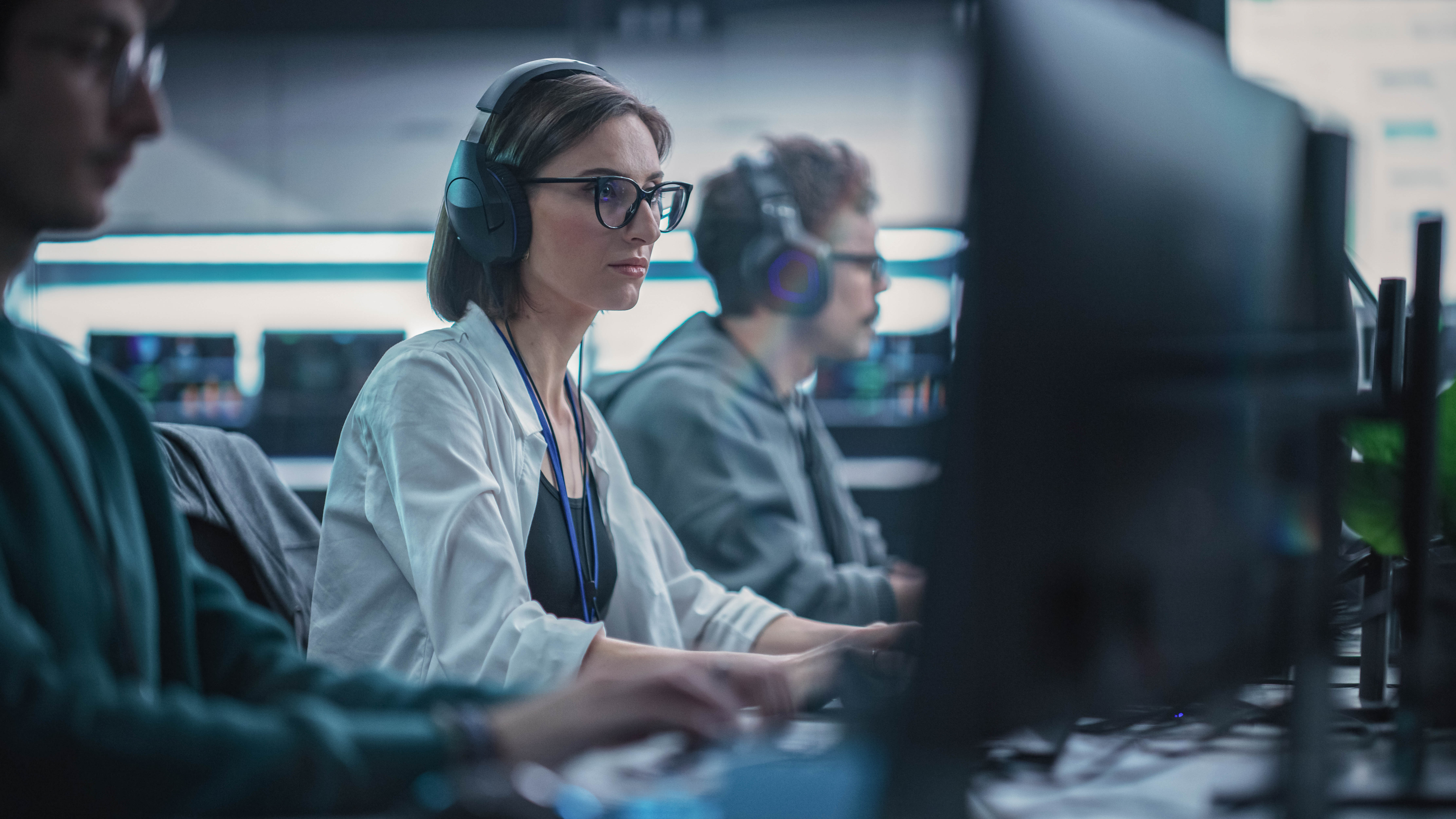 Drei Personen sitzen an Computern in einem modernen Arbeitsumfeld, eine trägt Kopfhörer und schaut konzentriert auf den Bildschirm.