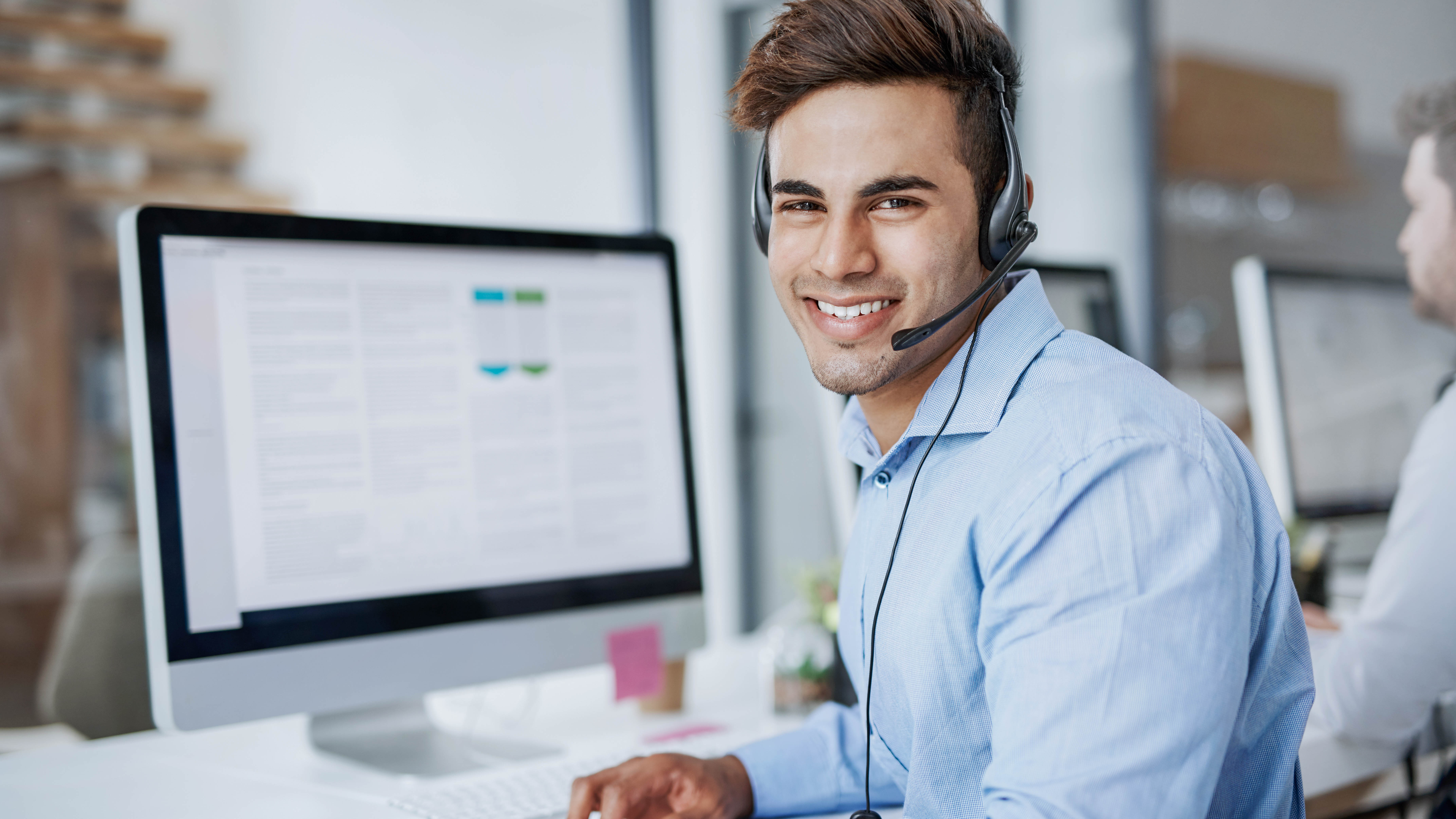 Lächelnder Mann mit Headset sitzt an einem Schreibtisch vor einem Computerbildschirm.