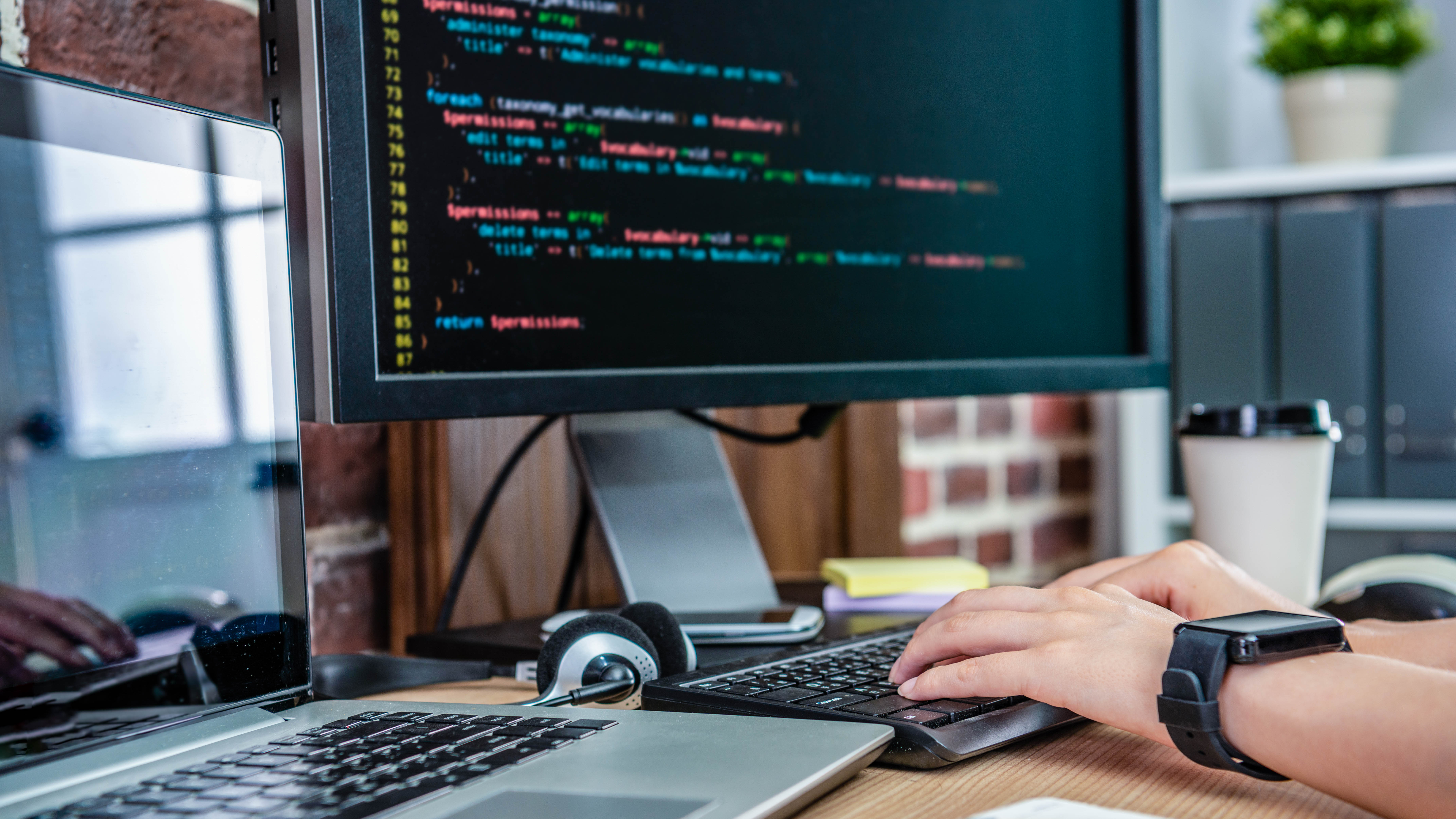 Eine Hand tippt auf einer Tastatur, während ein Monitor mit Programmiercode im Hintergrund sichtbar ist.
