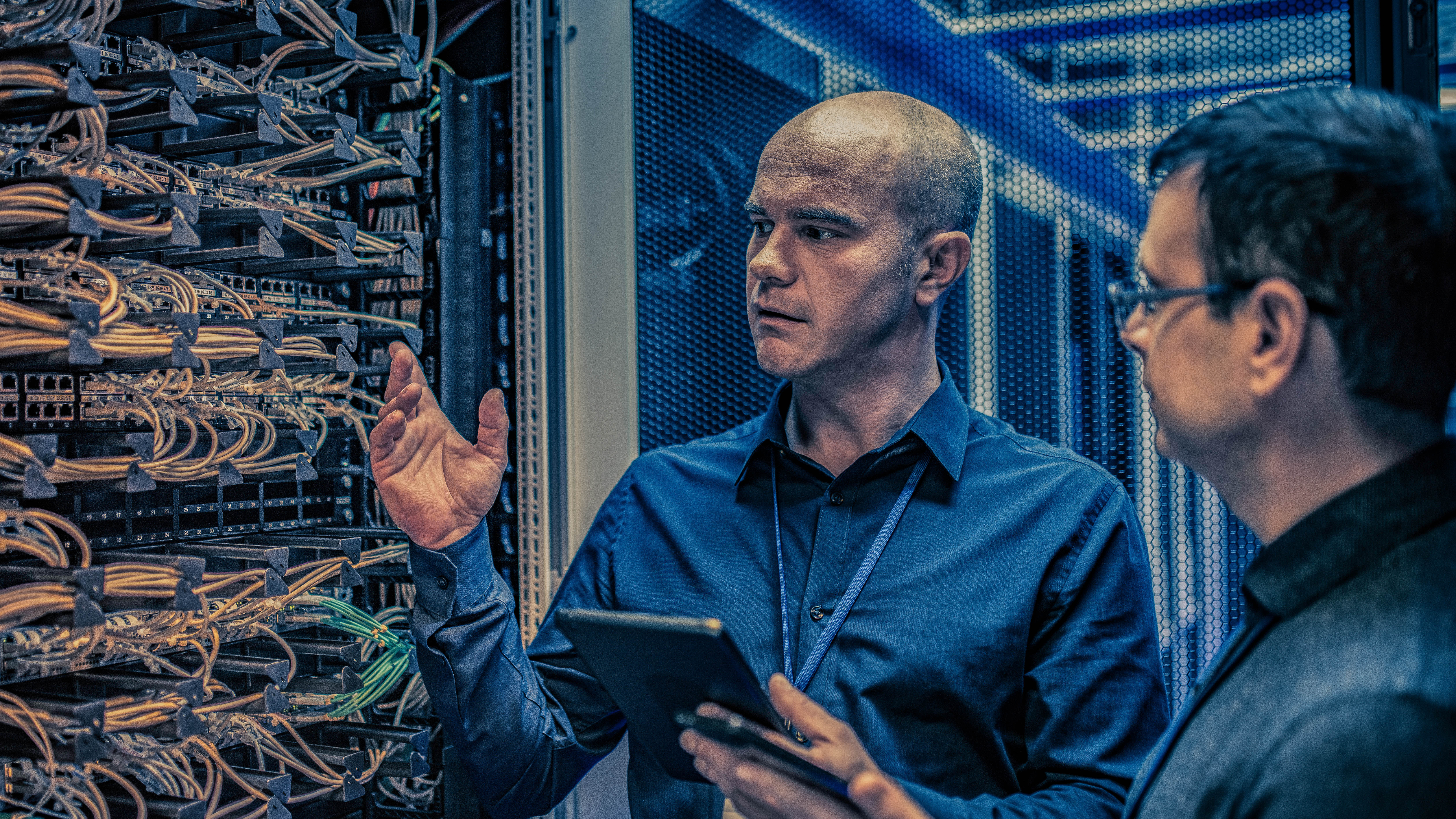 Zwei Männer stehen vor einem Serverrack mit Kabeln und diskutieren, während einer ein Tablet in der Hand hält.