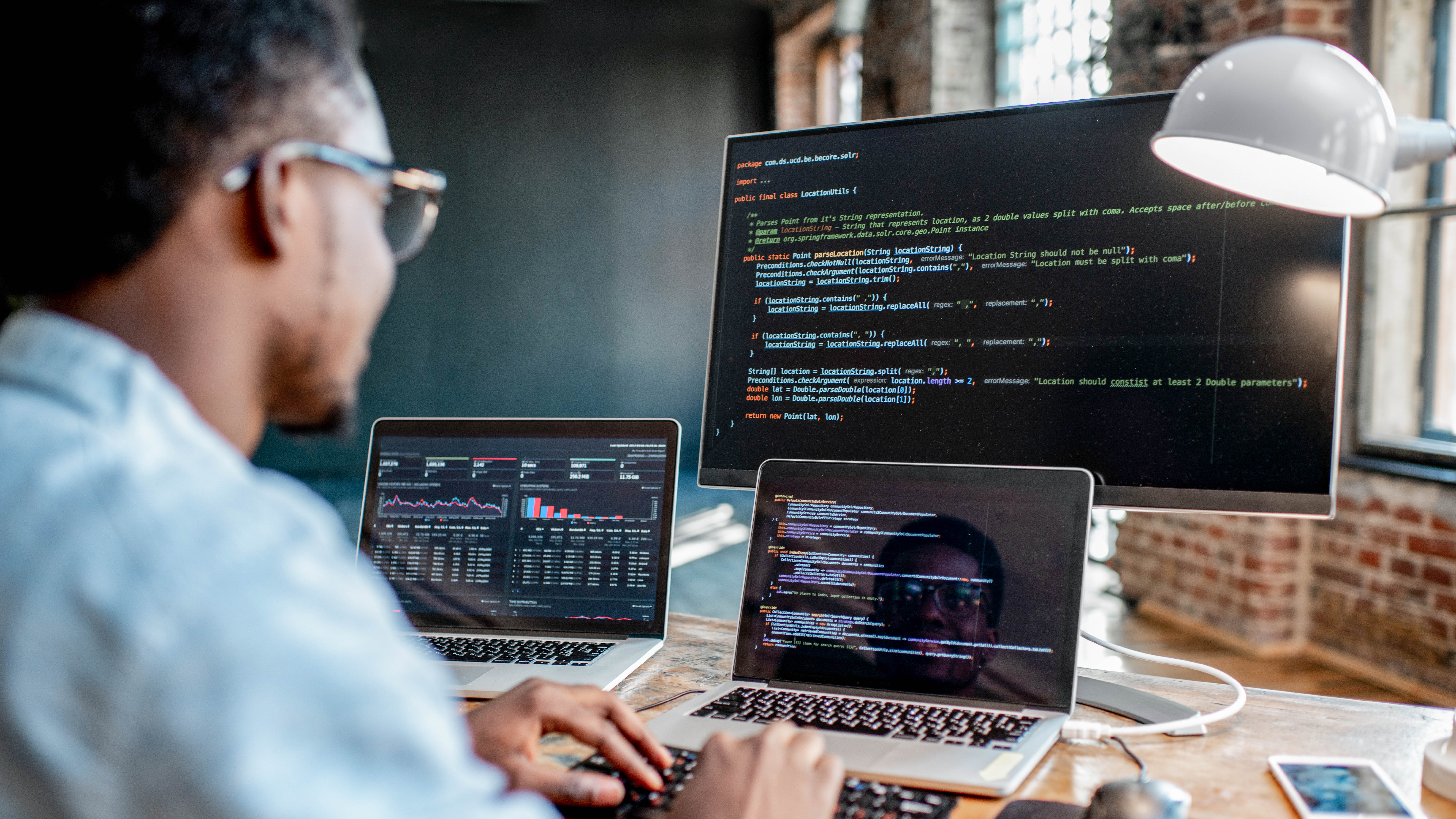 Ein Mensch arbeitet an zwei Laptops und einem Monitor, auf denen Programmiercode und Datenanalysen angezeigt werden.