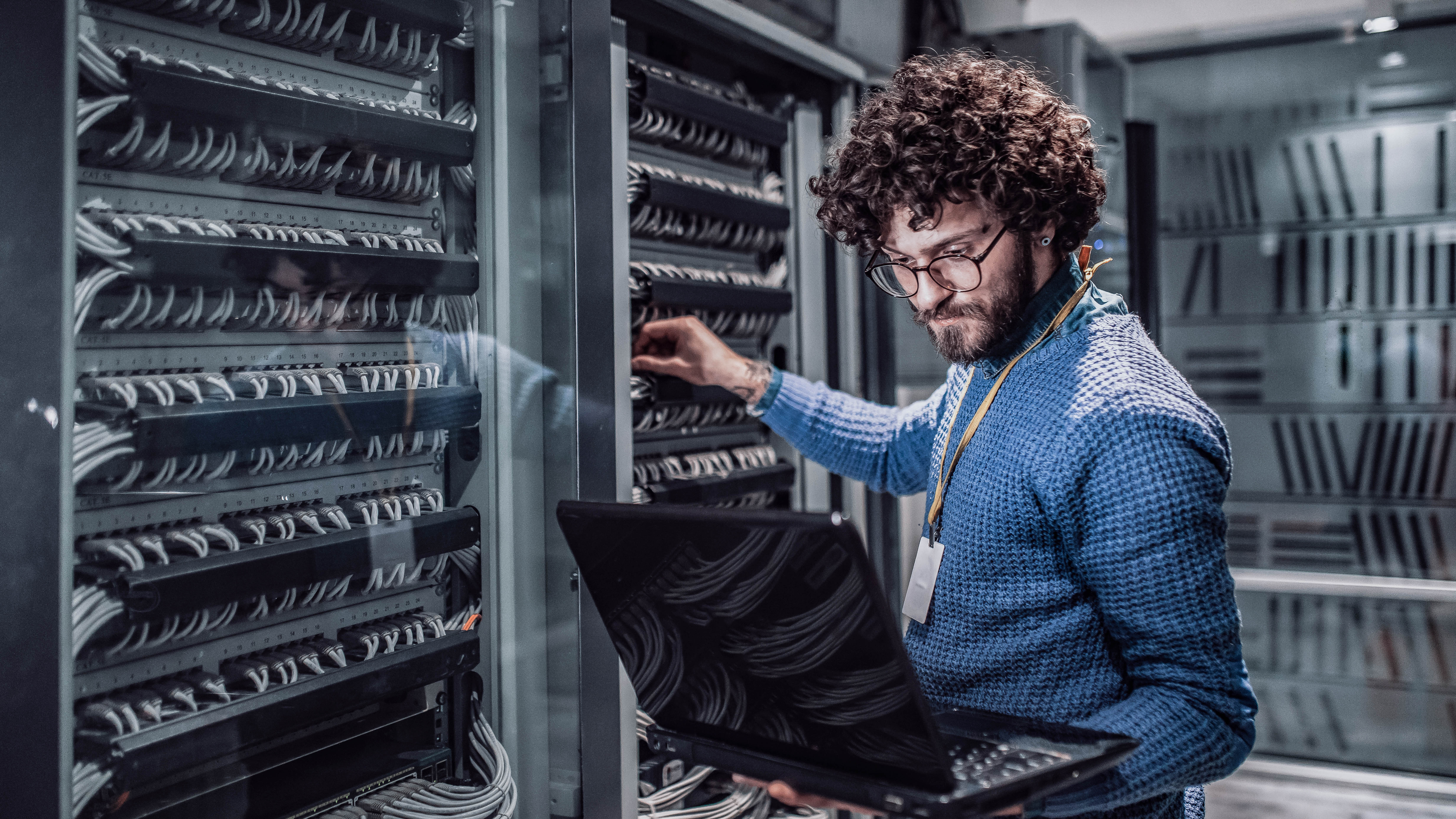Mann mit lockigem Haar arbeitet an einem Serverrack und hält einen Laptop in der Hand.