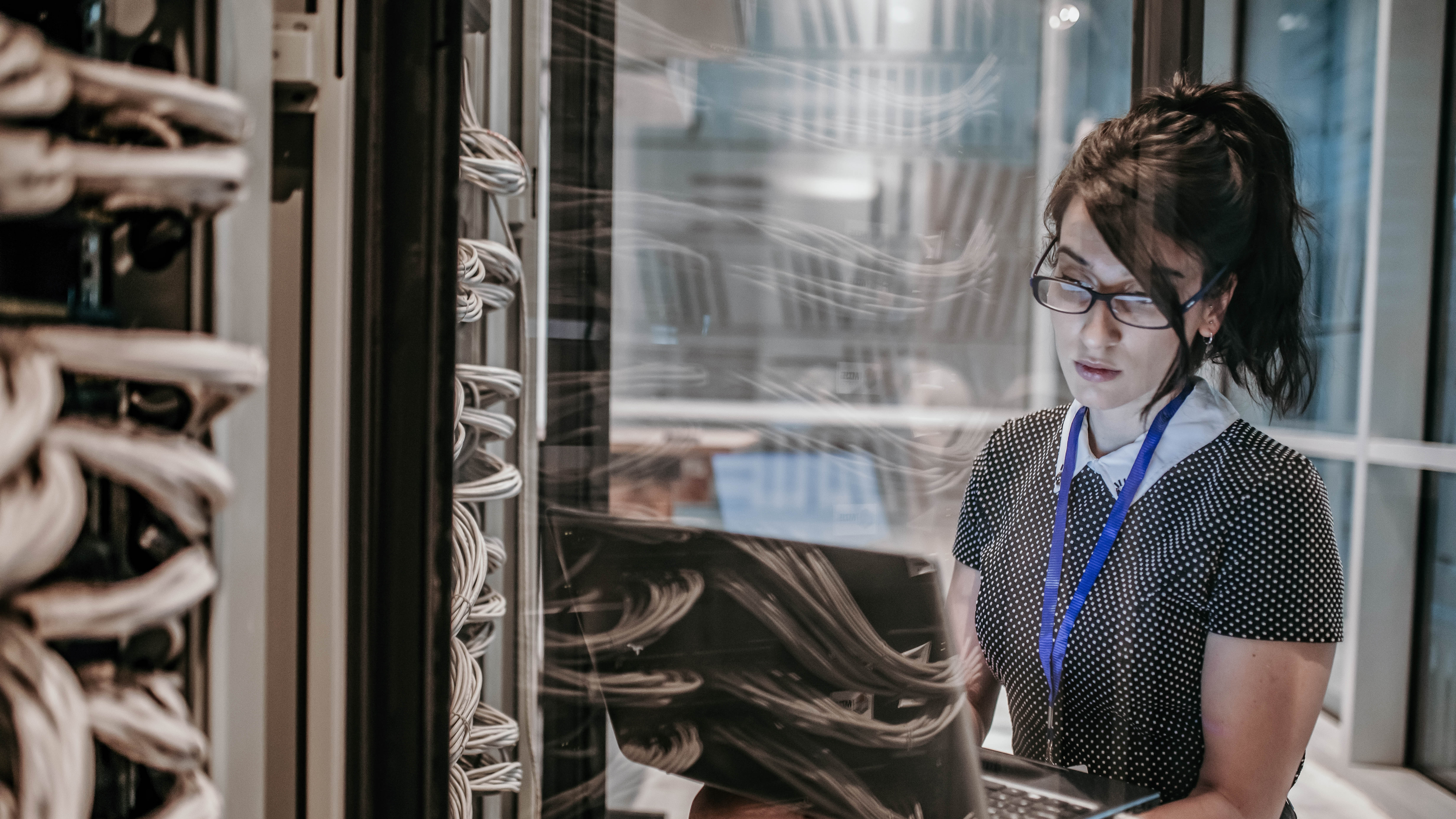 Frau mit Brille arbeitet an einem Laptop in einem Serverraum mit Kabeln im Hintergrund.