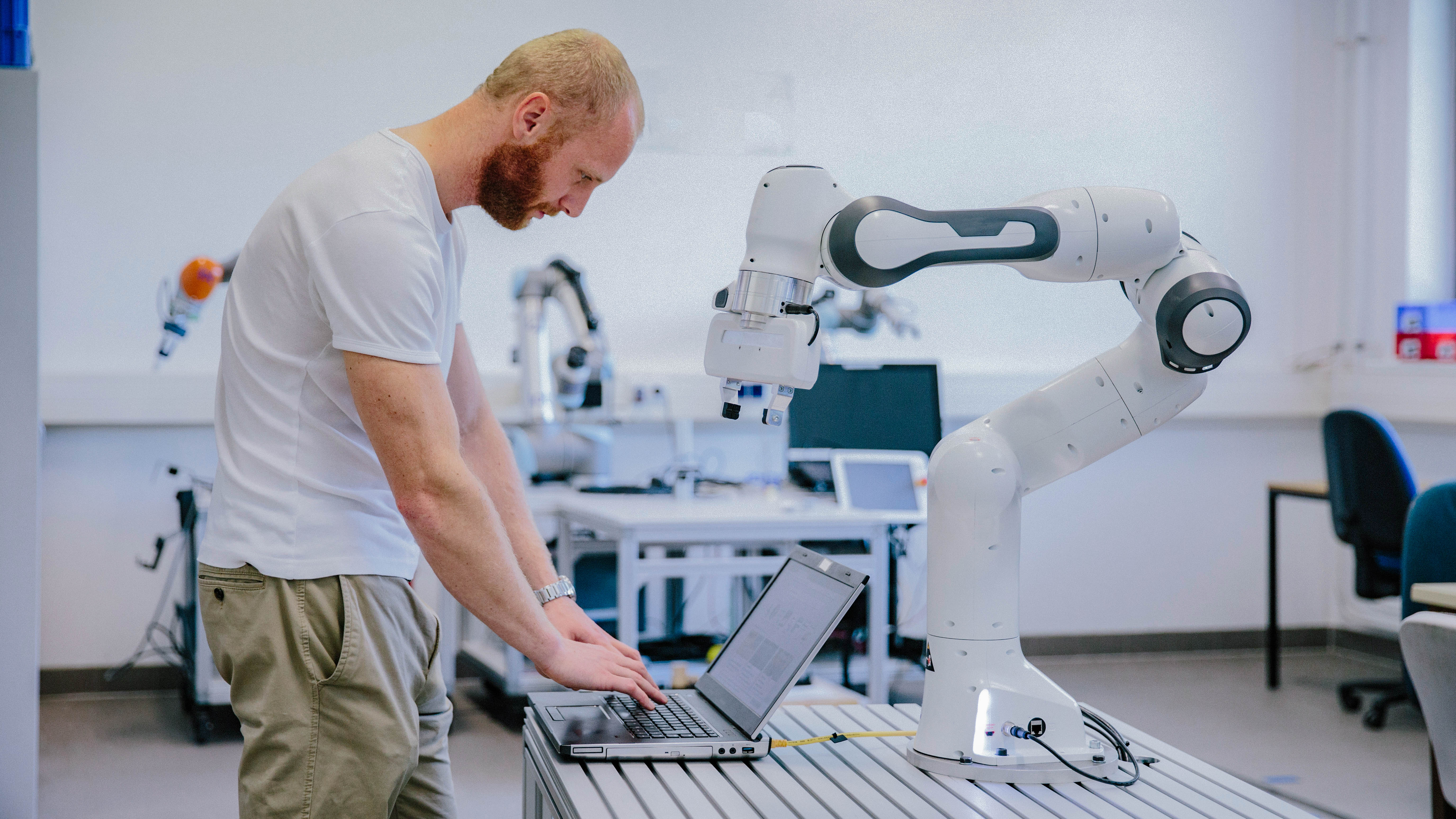 Ein Mann steht an einem Tisch und arbeitet mit einem Laptop, während ein Roboterarm neben ihm positioniert ist.