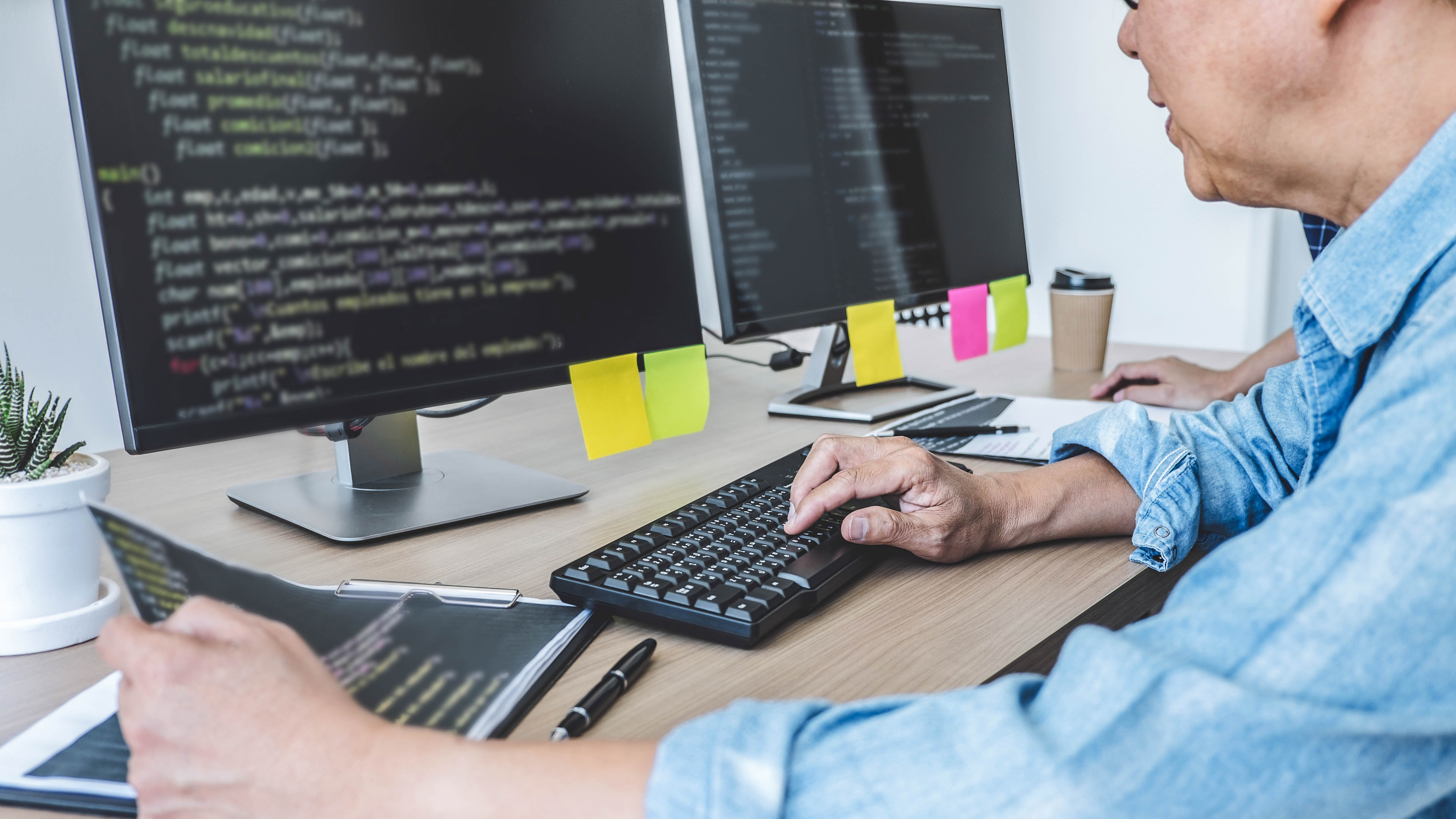 Zwei Personen arbeiten an Computern mit Programmiercode auf den Bildschirmen, Notizen und eine Kaffeetasse sind sichtbar.