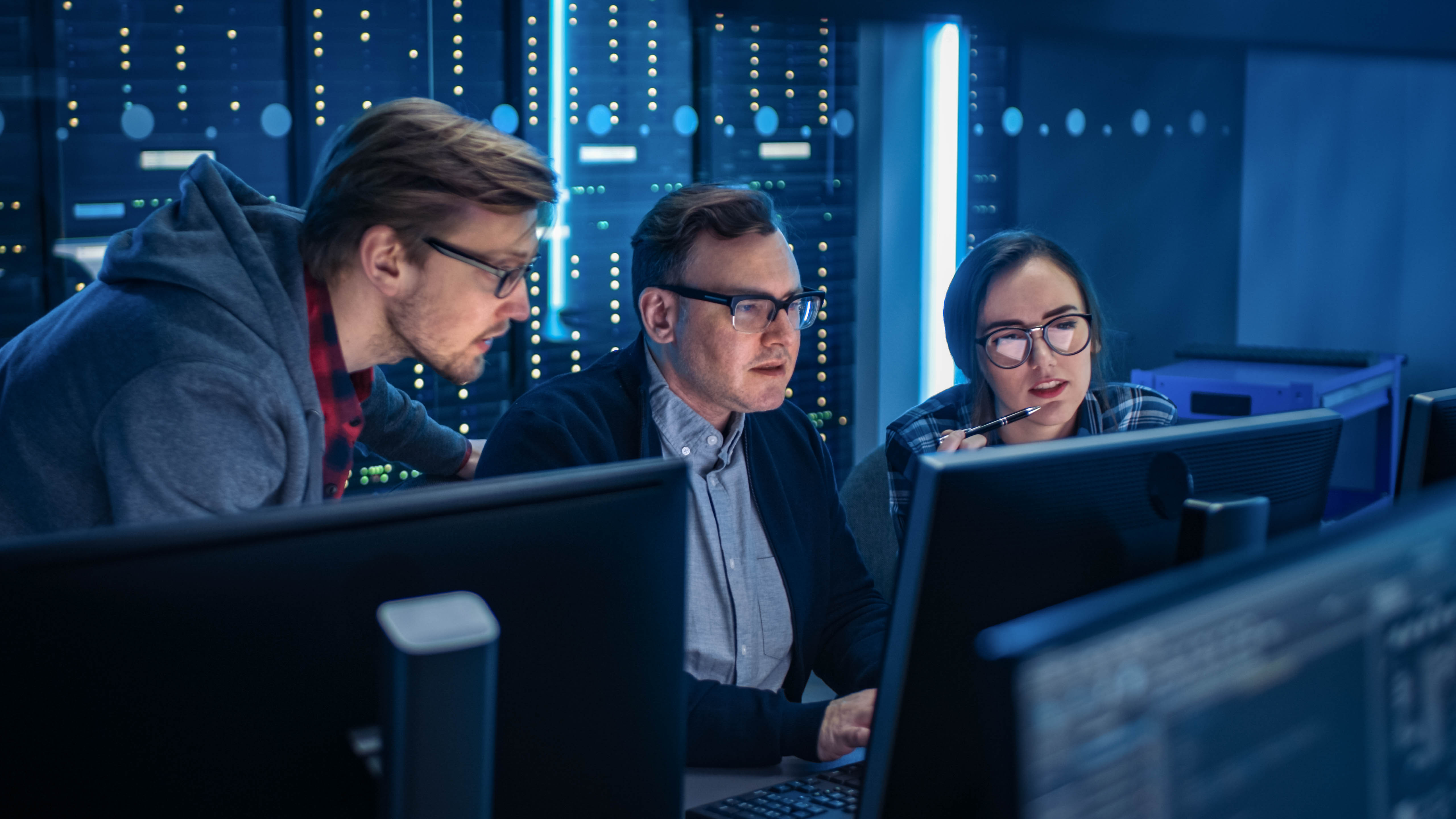 Drei Personen arbeiten konzentriert an Computern in einem Serverraum mit blauen Lichtern und Server-Racks im Hintergrund.