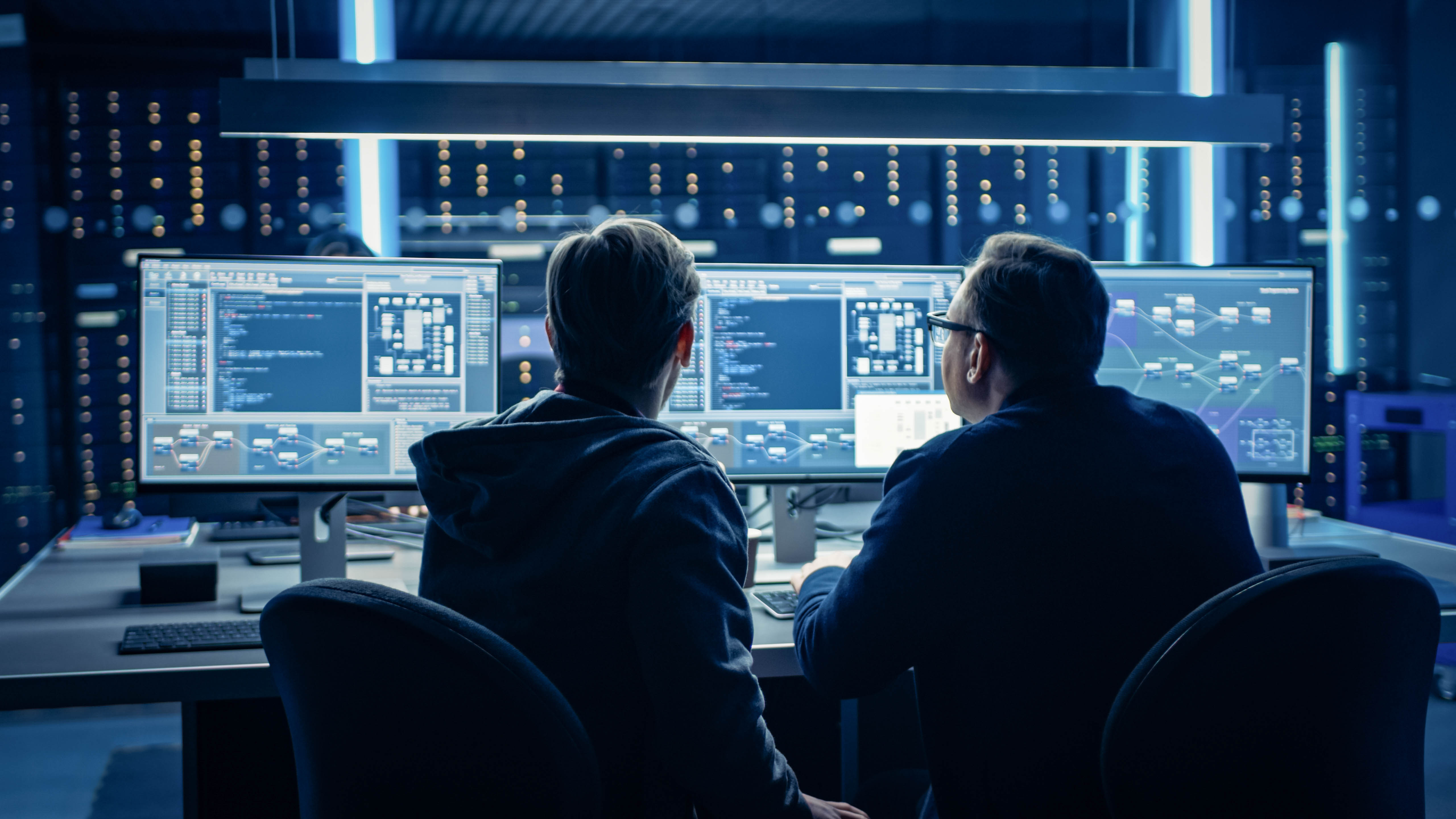 Zwei Personen sitzen an einem Tisch vor mehreren Monitoren mit Programmiercode und Diagrammen in einem dunklen Raum.