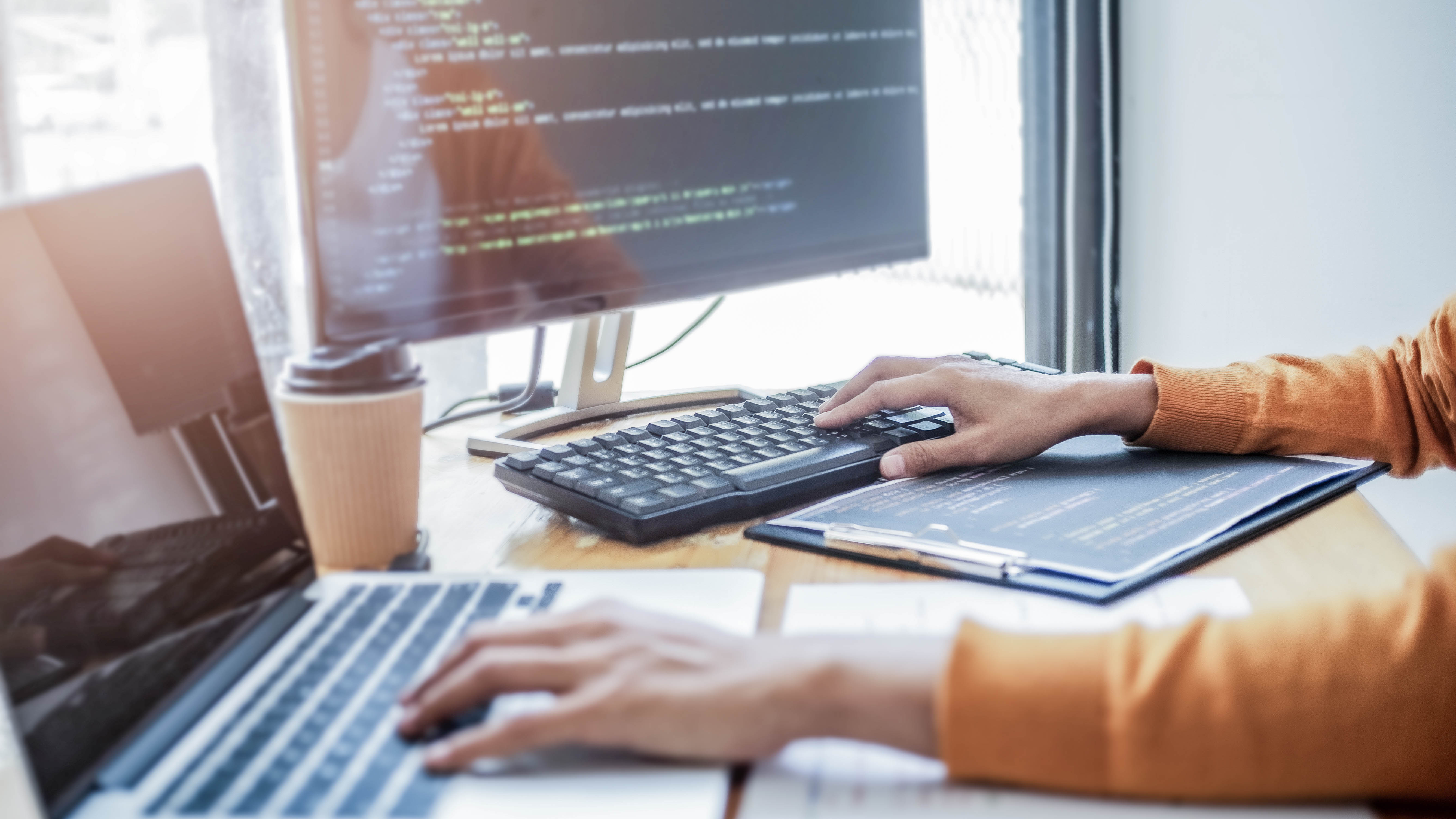 Hände tippen auf einer Tastatur, während ein Laptop und ein Monitor mit Code sichtbar sind. Ein Kaffeebecher steht auf dem Tisch.