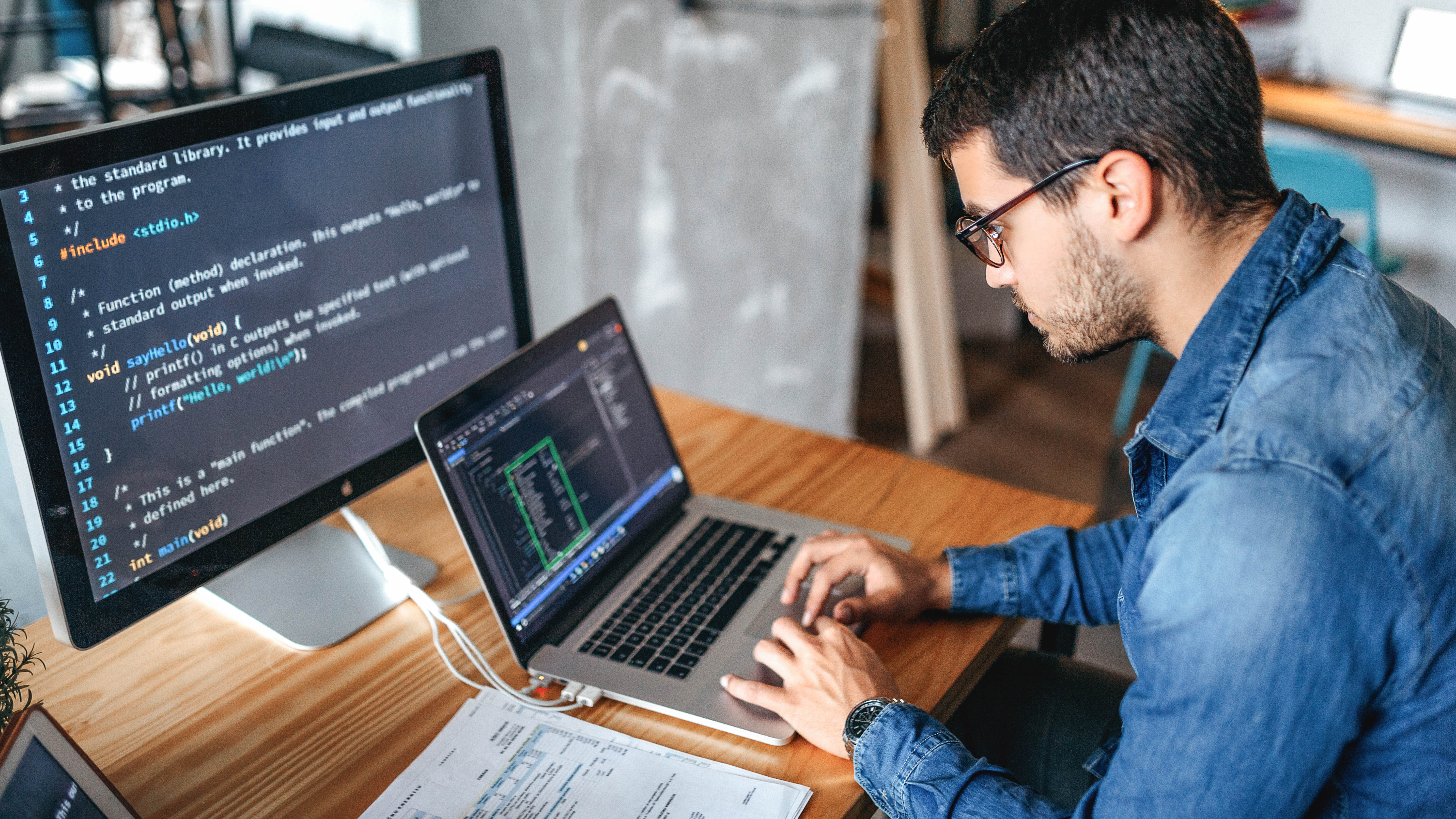 Ein Mann sitzt an einem Tisch und programmiert auf einem Laptop, während ein großer Monitor Code anzeigt.