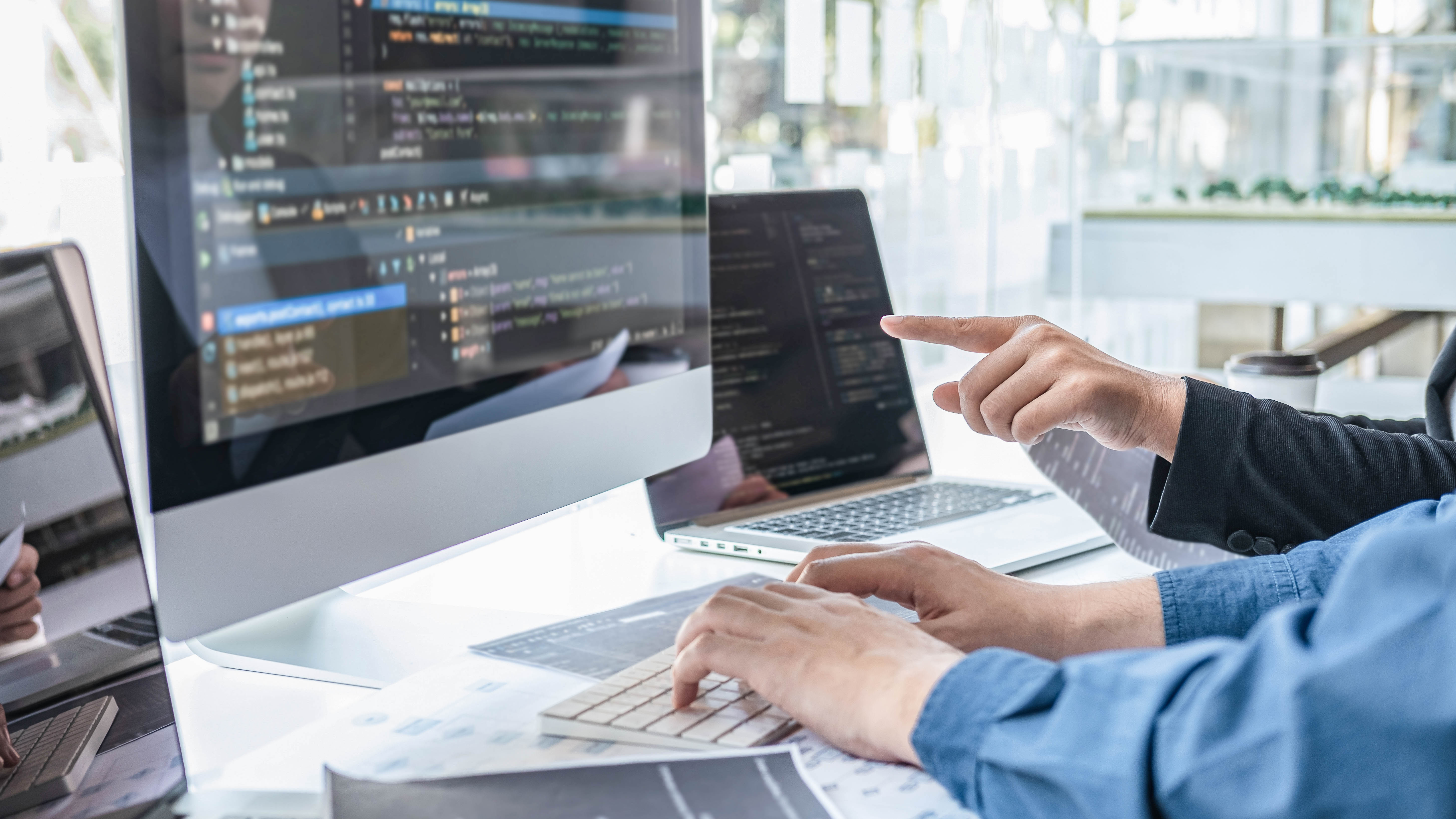 Hände von zwei Personen, die an einem Computer arbeiten, während auf dem Bildschirm Programmiercode angezeigt wird.