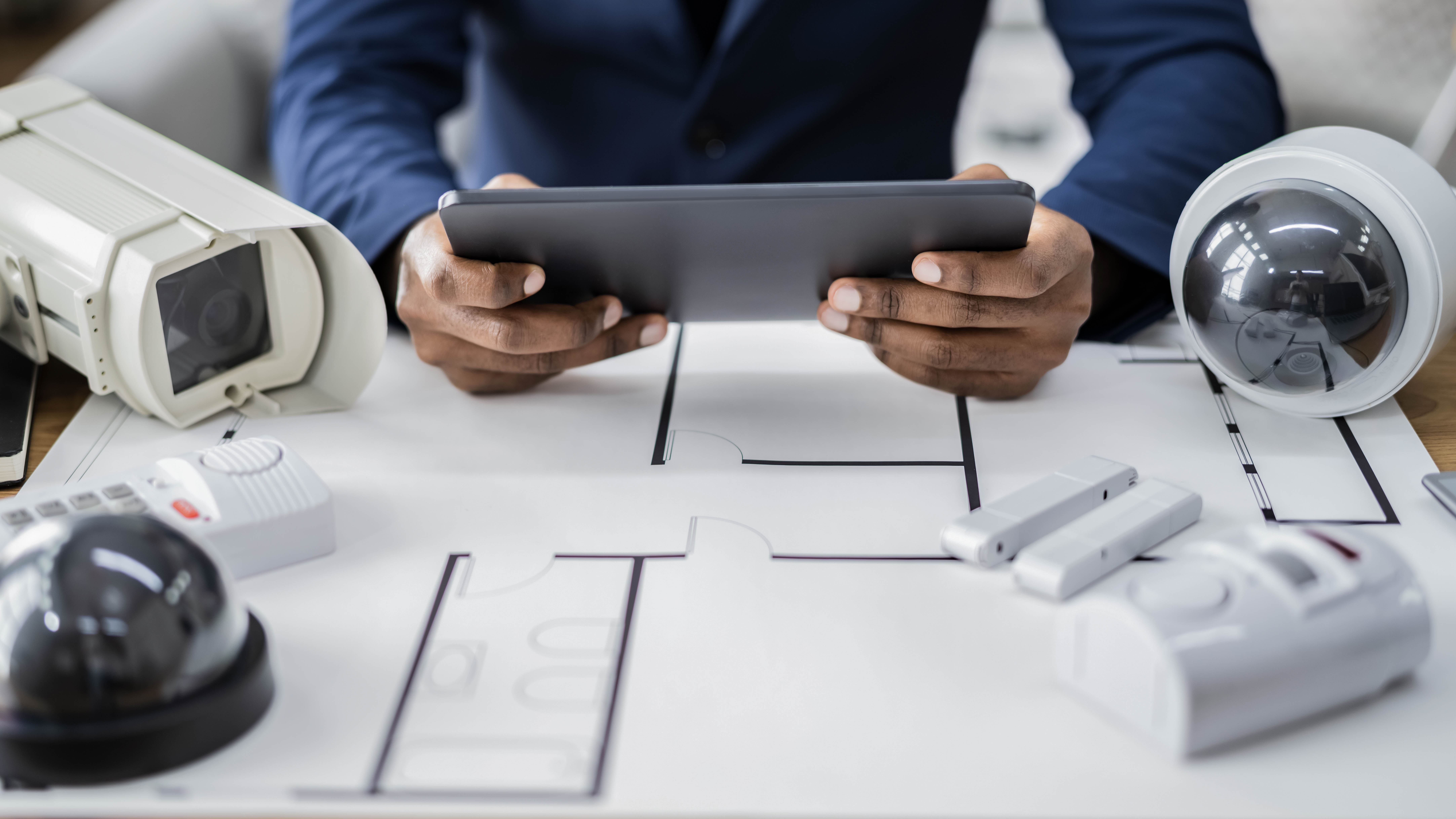 Hände halten ein Tablet über einem Plan, umgeben von Sicherheitskameras und Sensoren auf einem Tisch.