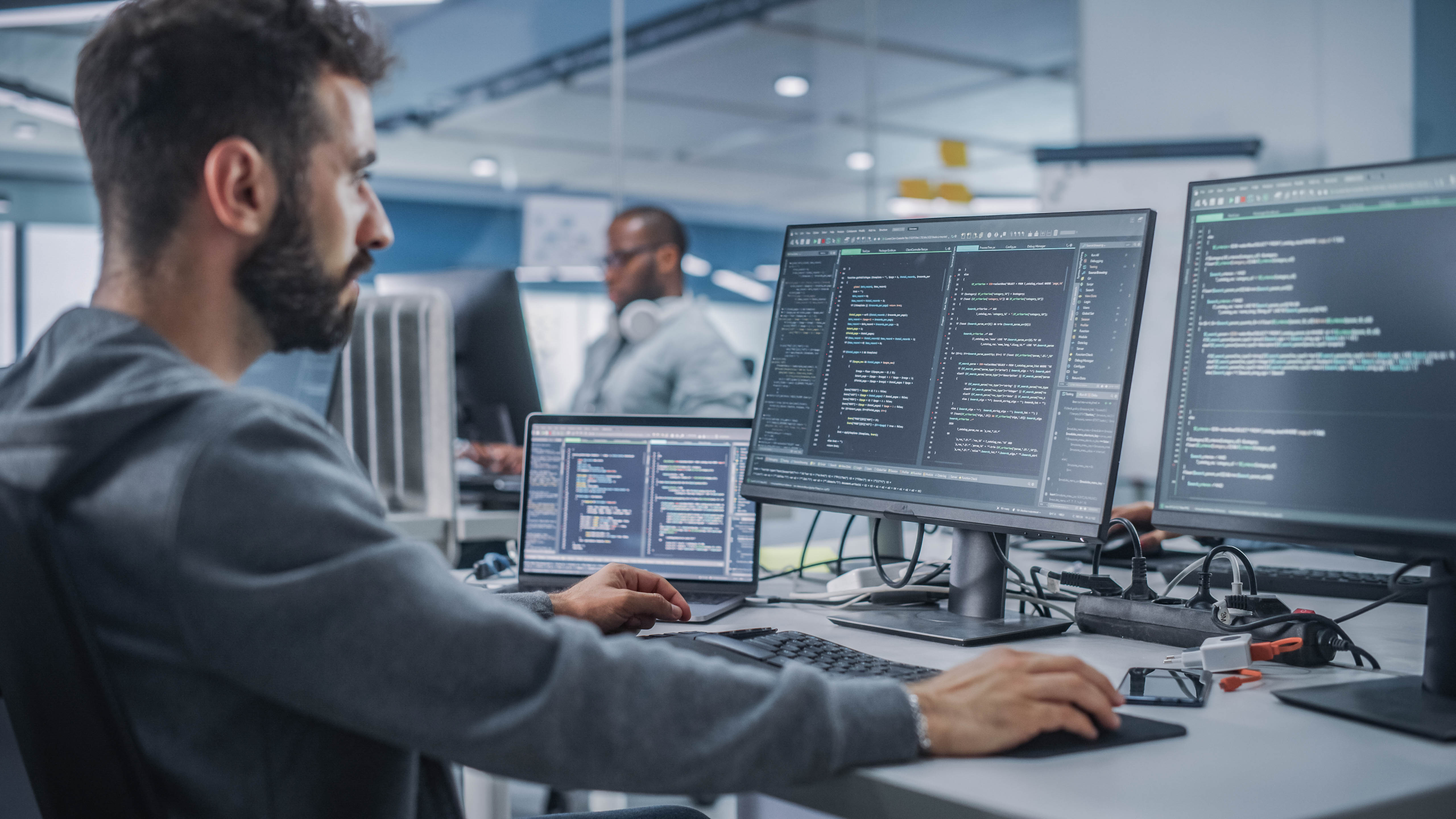 Mann arbeitet an mehreren Bildschirmen mit Programmiercode in einem modernen Büro.