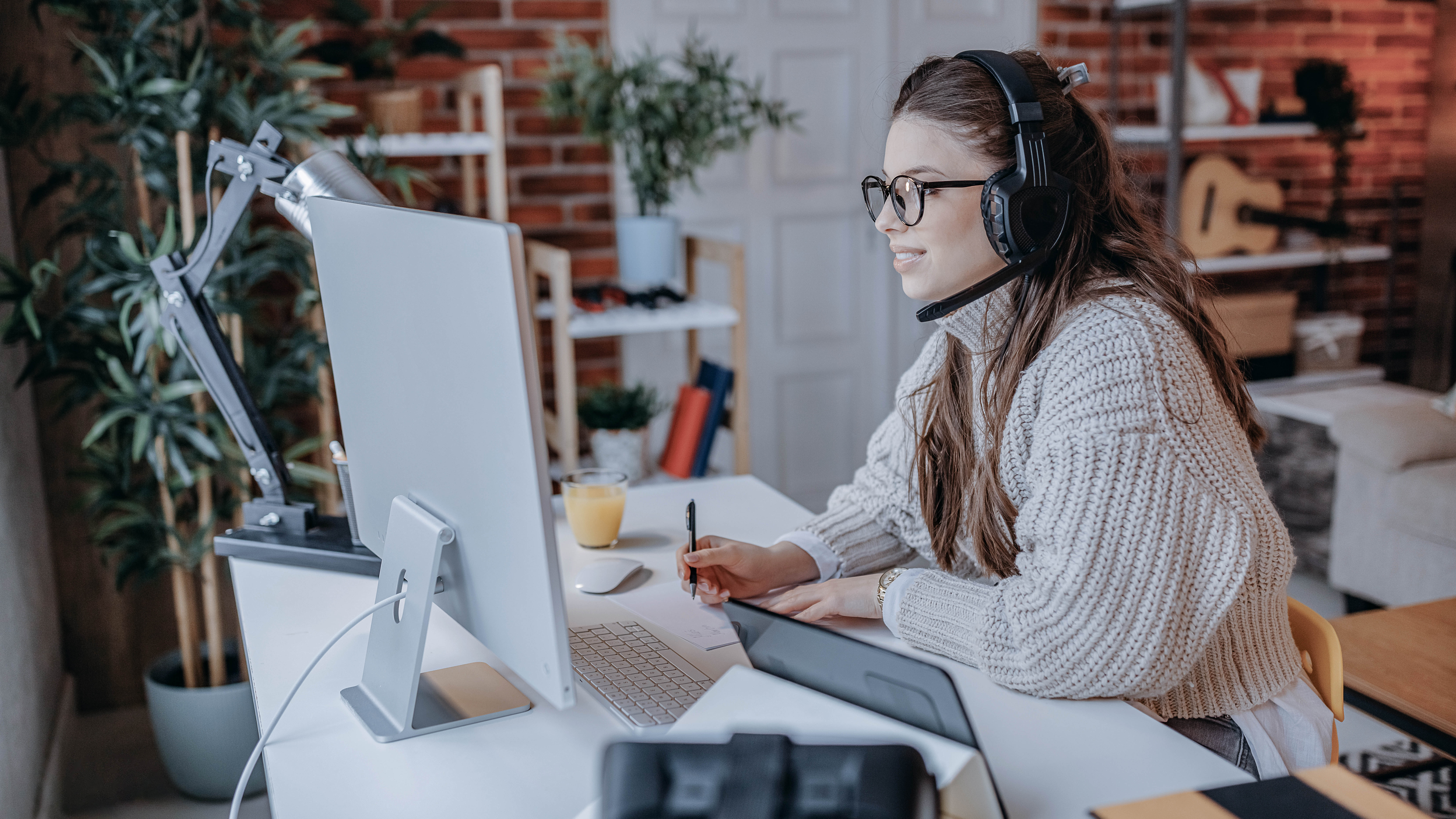Frau mit Headset sitzt an einem Schreibtisch, arbeitet am Computer und schreibt Notizen.