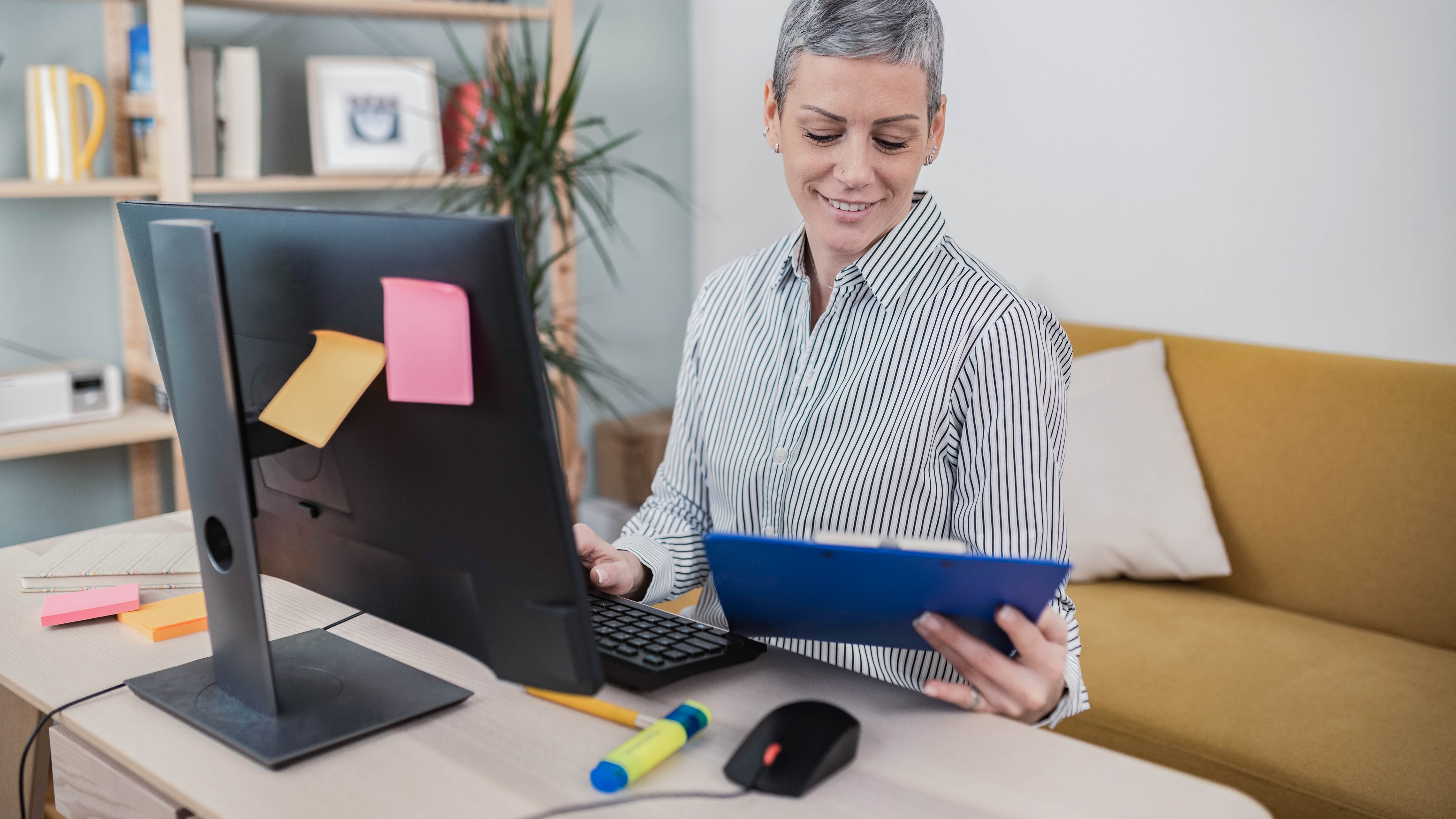 Eine Person mit kurzen grauen Haaren sitzt an einem Schreibtisch und arbeitet am Computer, während sie ein blaues Dokument in der Hand hält.