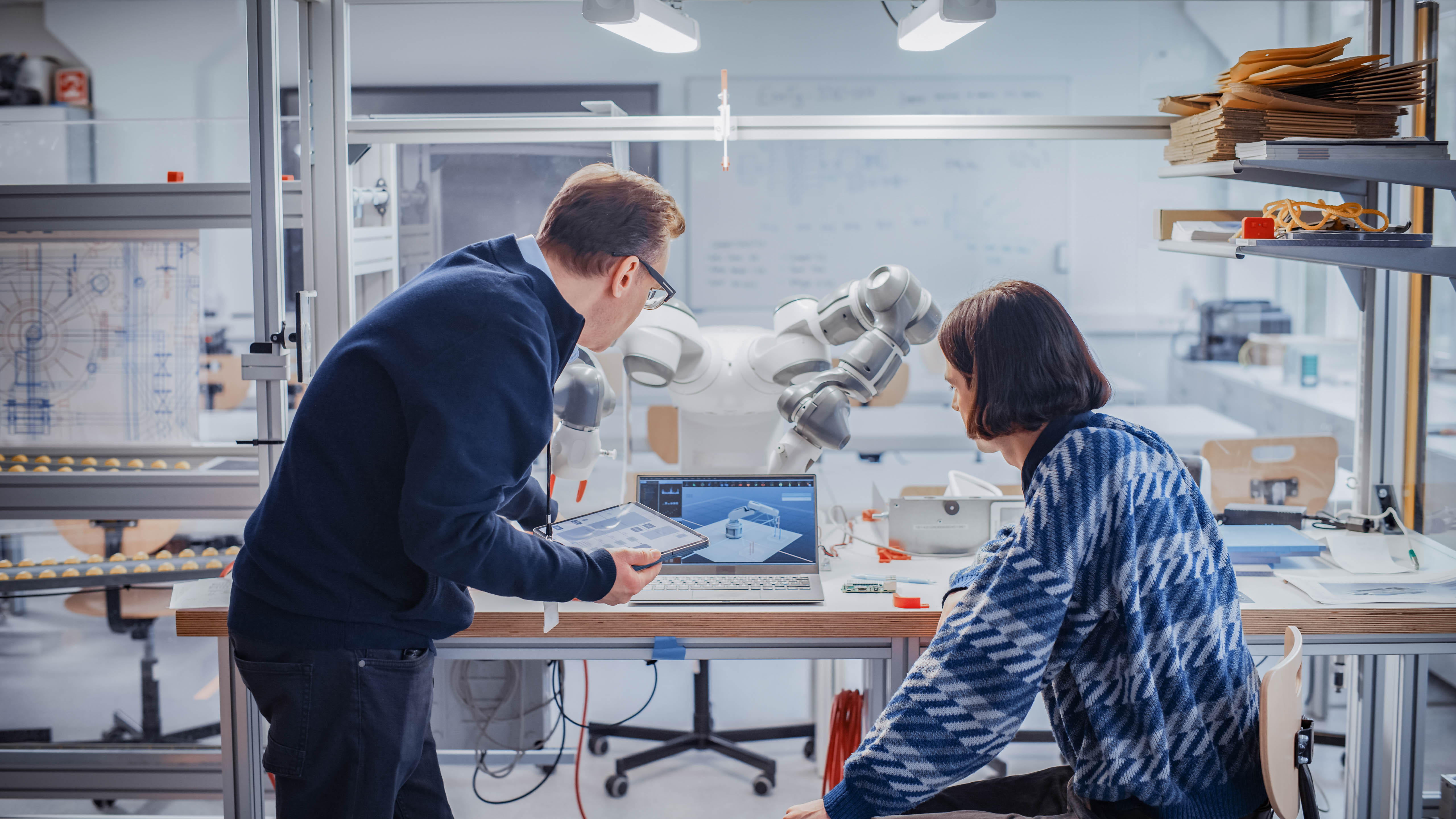 Zwei Personen arbeiten an einem Tisch mit einem Laptop und einem Tablet, während ein Roboterarm im Hintergrund sichtbar ist.