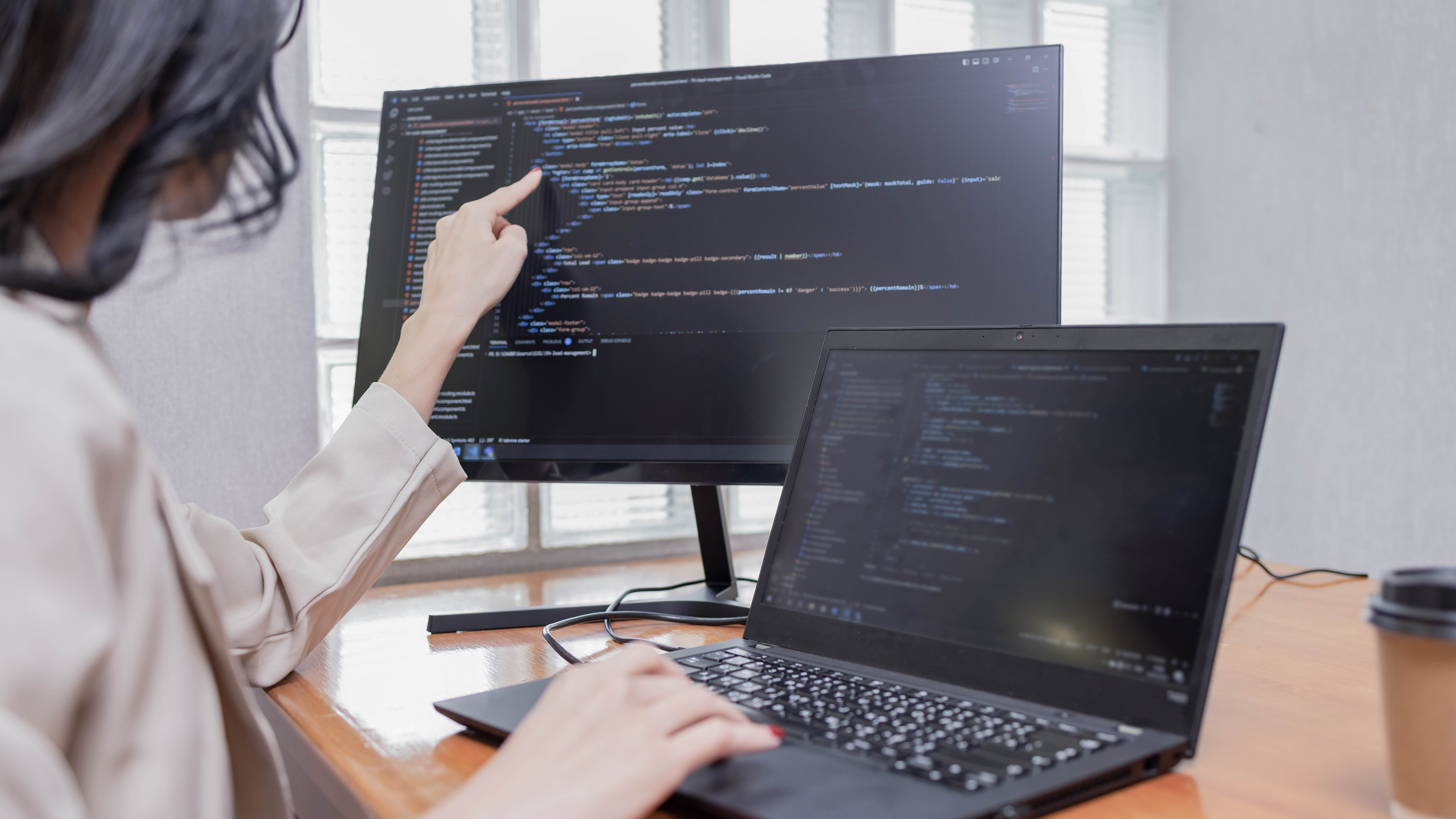 Eine Person zeigt mit dem Finger auf den Bildschirm eines Monitors, während sie an einem Laptop tippt, beide zeigen Programmiercode.