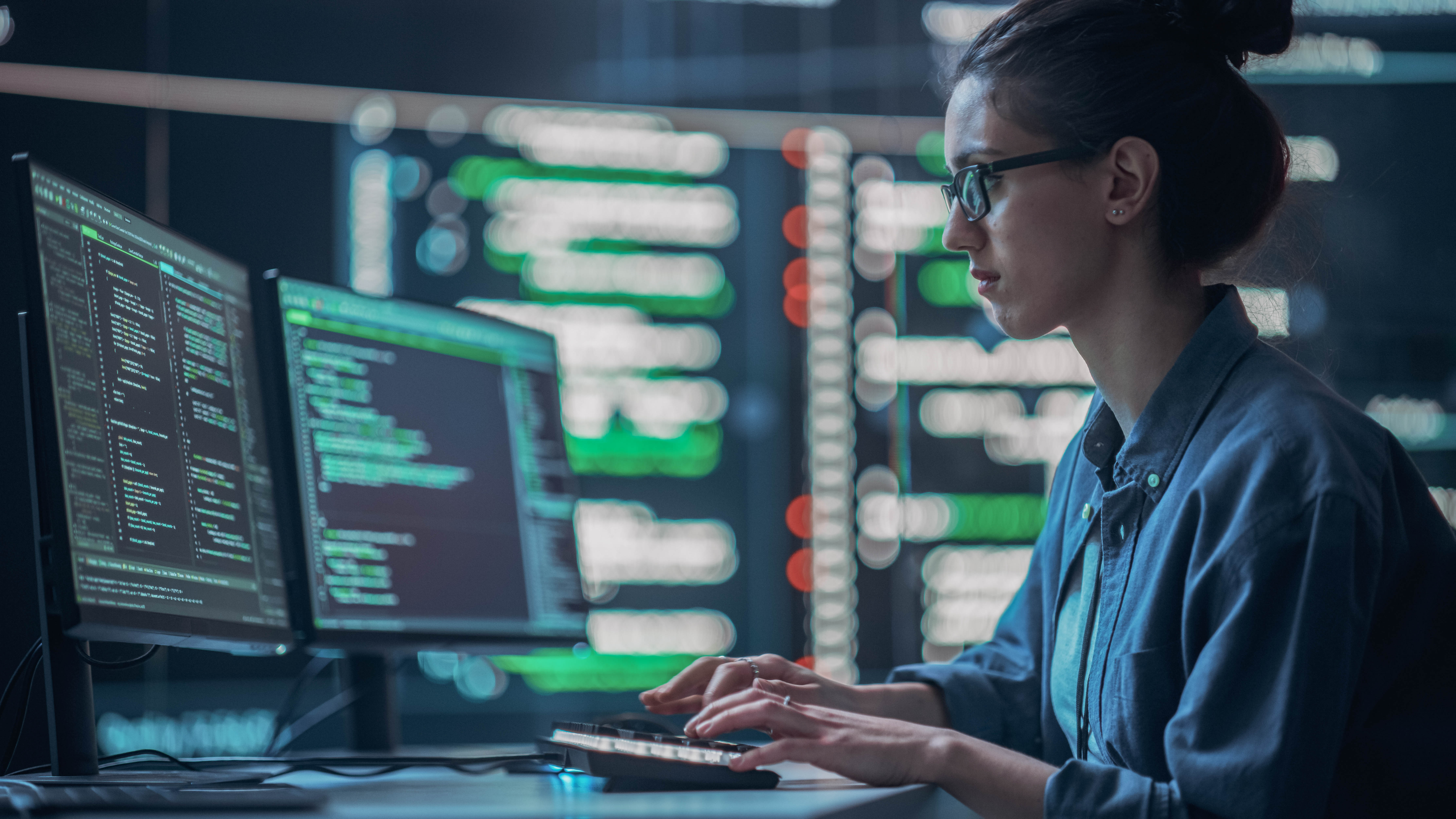 Eine Person mit Brille sitzt an einem Tisch und tippt auf einer Tastatur, während sie auf zwei Computerbildschirme mit Code schaut.