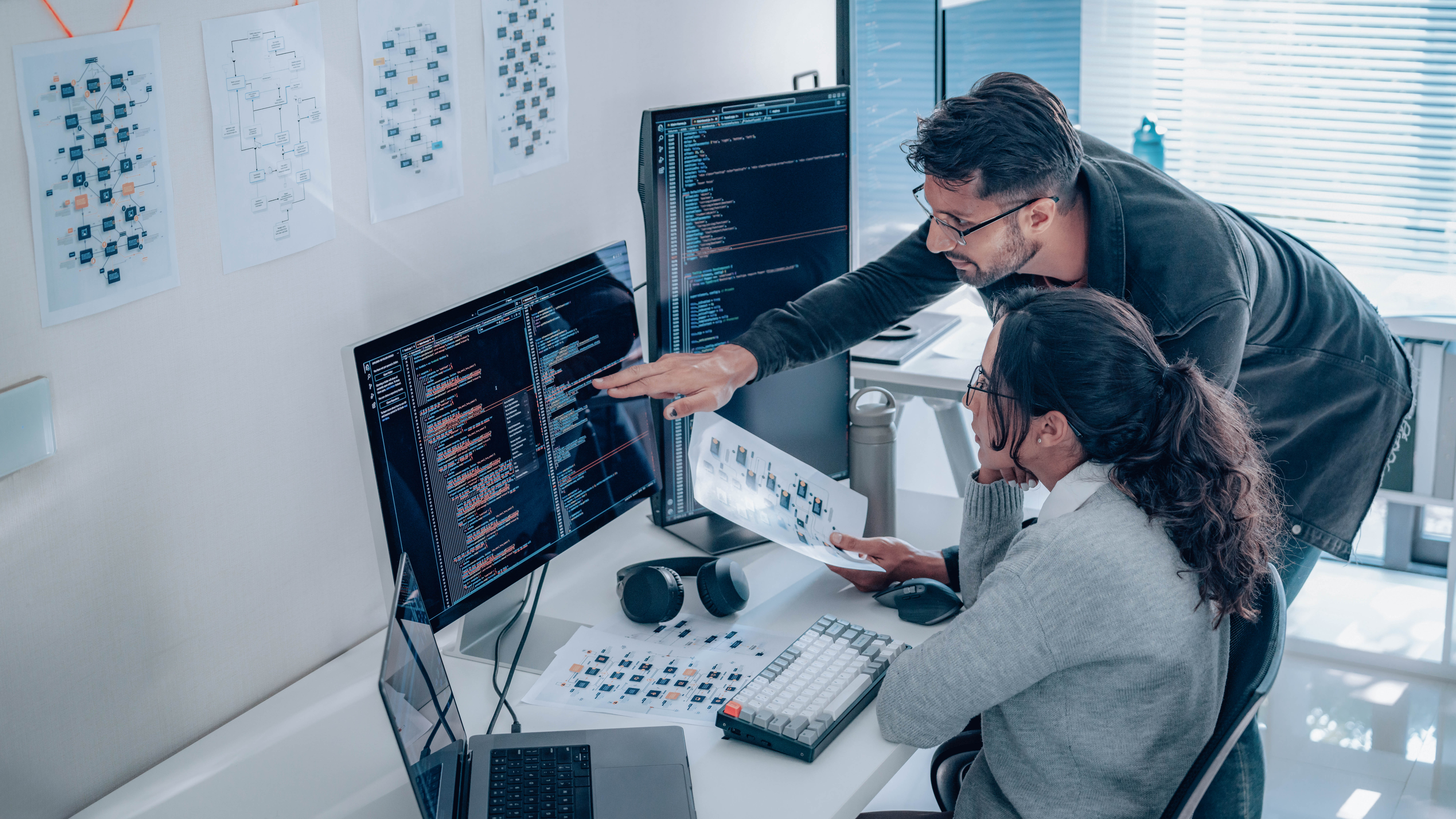 Zwei Personen arbeiten an einem Schreibtisch mit mehreren Monitoren, auf denen Code und Diagramme angezeigt werden.