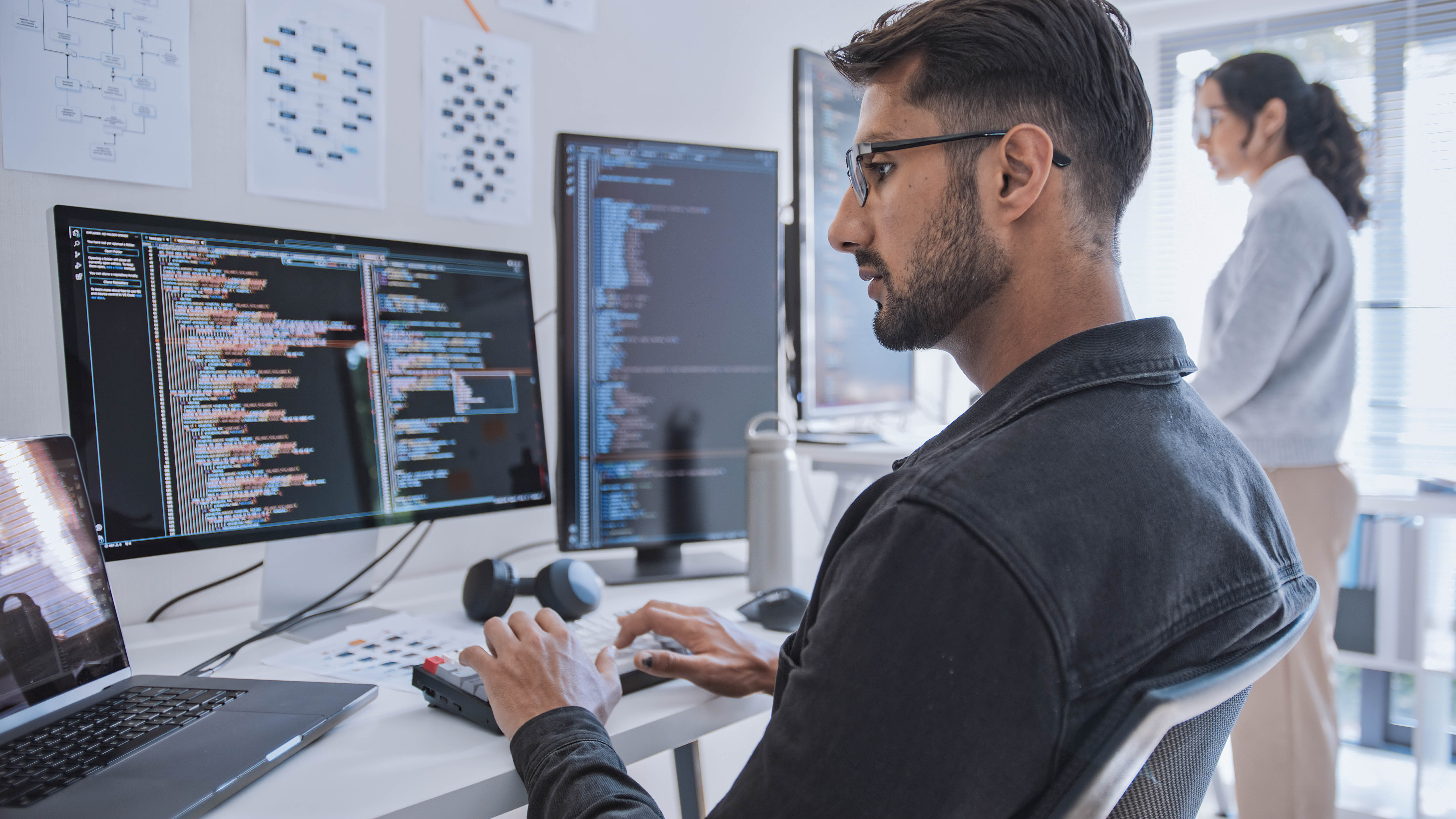 Ein Mann sitzt an einem Schreibtisch und arbeitet an mehreren Monitoren mit Programmiercode, während eine Frau im Hintergrund steht.