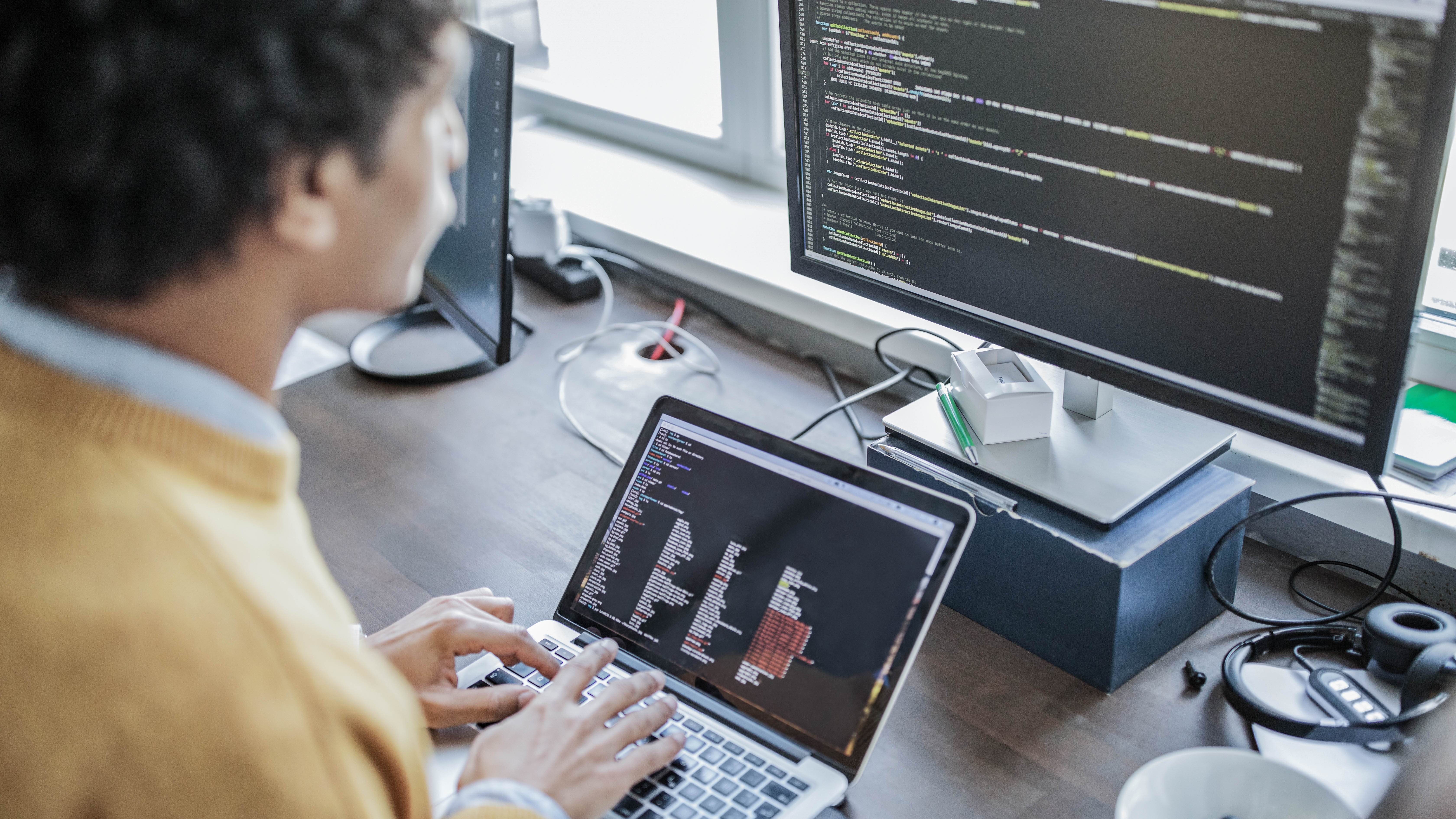 Person arbeitet an einem Laptop mit sichtbarem Code, während ein zweiter Monitor ebenfalls Code anzeigt.