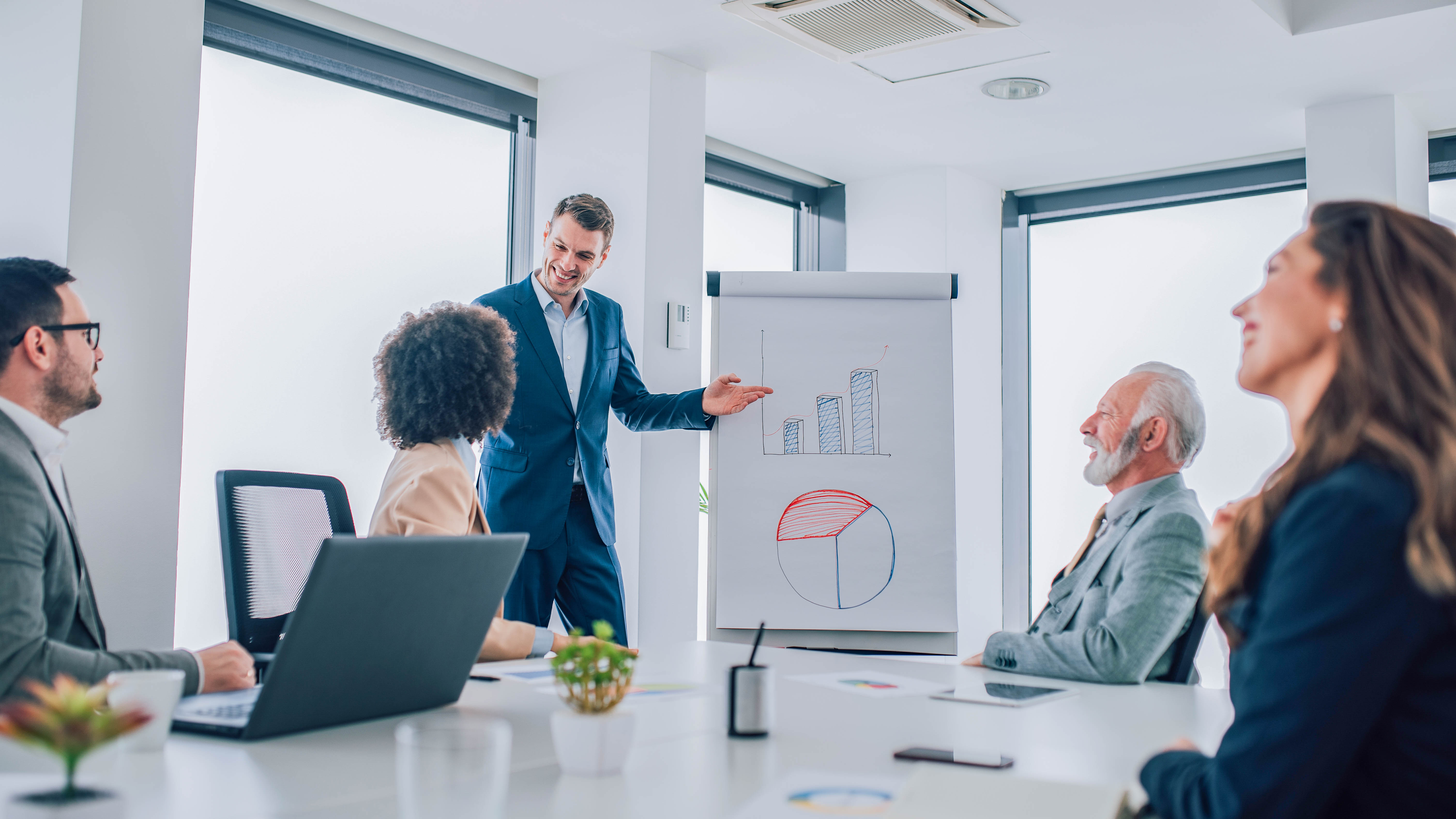 Ein Mann präsentiert vor einer Gruppe in einem Konferenzraum, während ein Flipchart mit Diagrammen im Hintergrund steht.