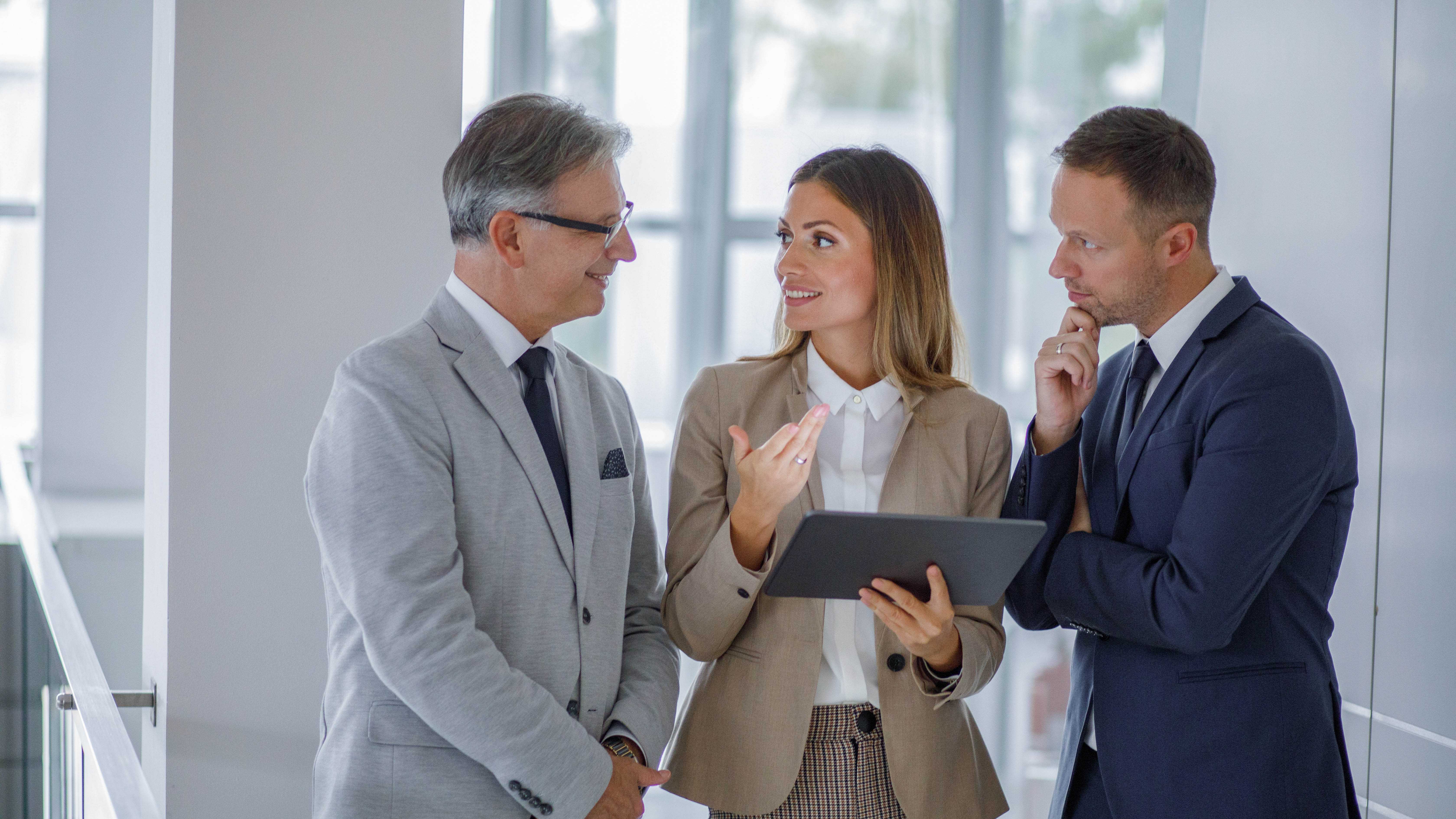 Drei Personen in Businesskleidung diskutieren in einem modernen Büro, eine Person hält ein Tablet.