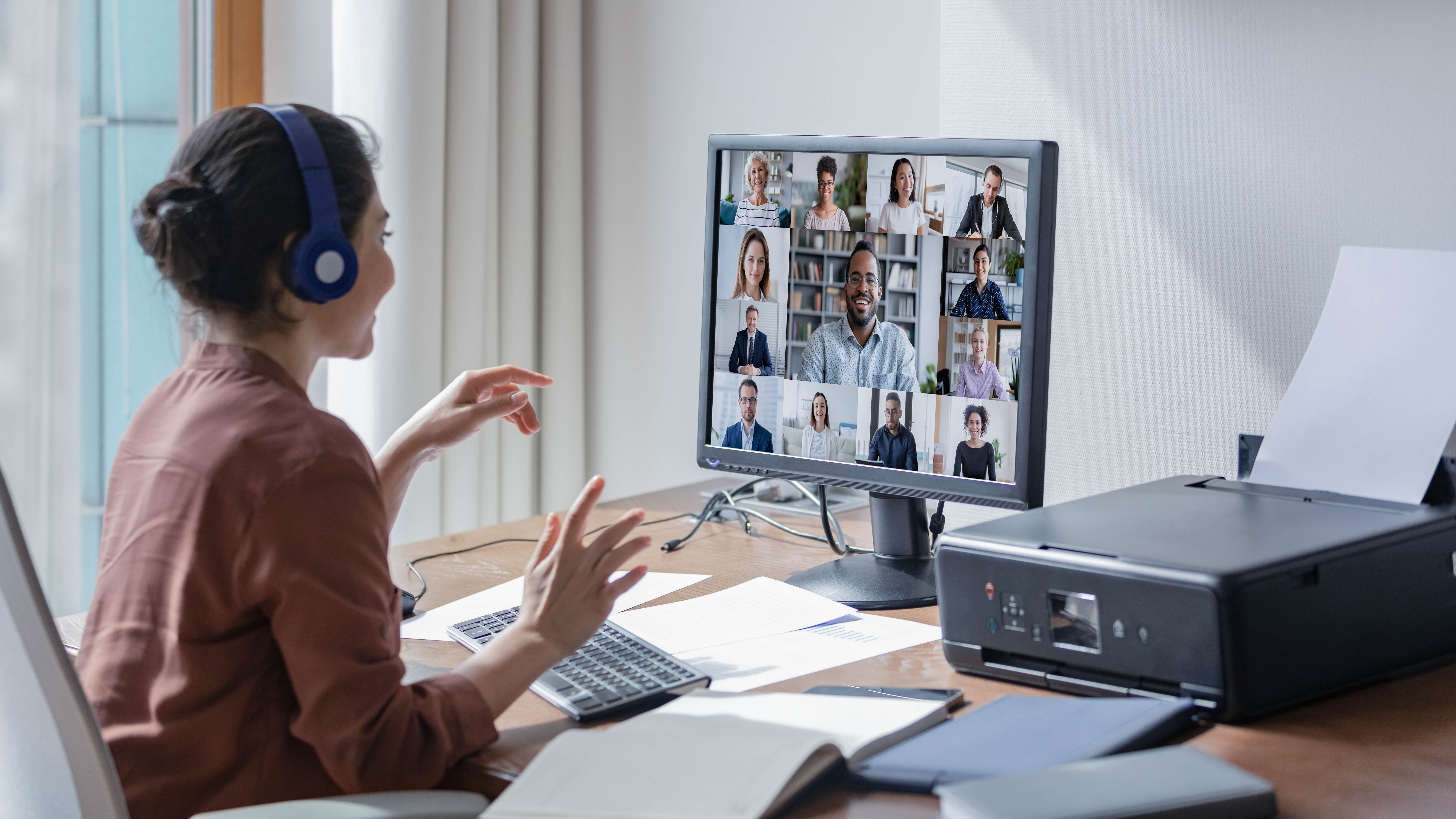 Frau mit Kopfhörern interagiert während einer Videokonferenz mit mehreren Teilnehmern auf einem Monitor.