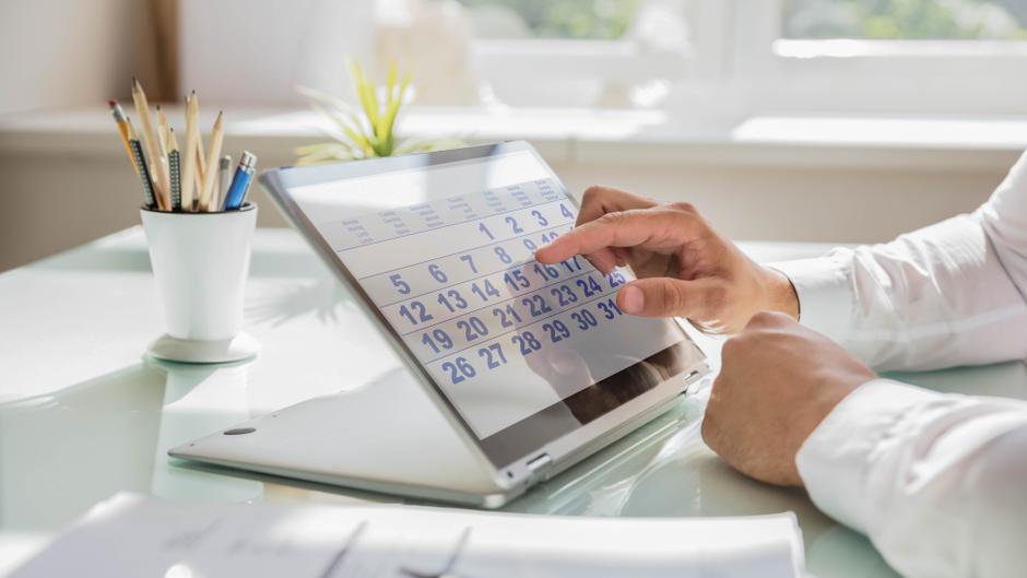 Eine Hand tippt auf einen digitalen Kalender auf einem Tablet, daneben ein Topf mit Stiften.