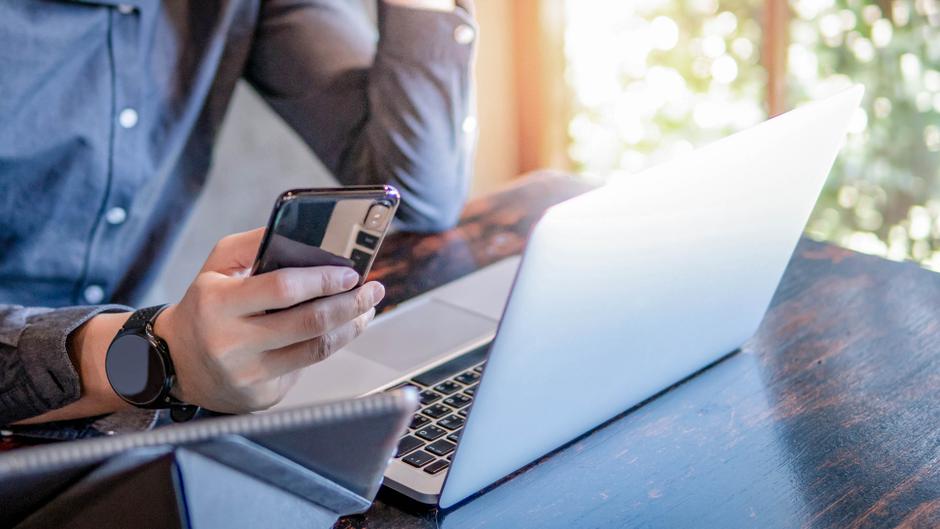 Eine Person hält ein Smartphone in der Hand, während ein Laptop auf dem Tisch steht.