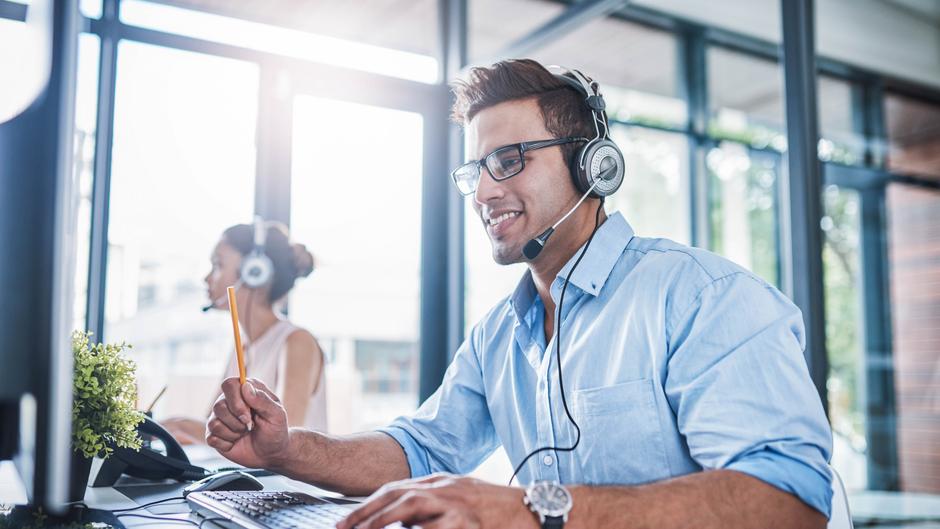 Mann mit Headset lächelt am Computer, während eine Frau im Hintergrund ebenfalls telefoniert.