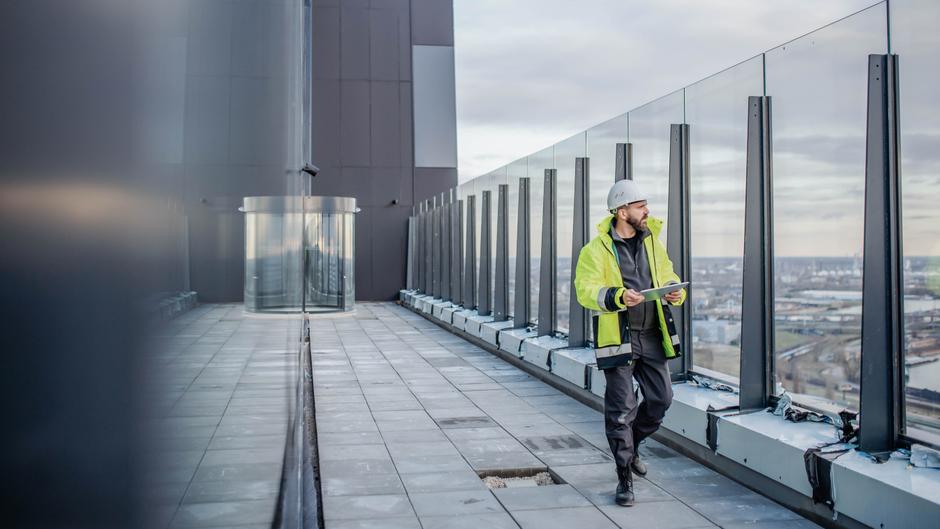 Ein Bauarbeiter in Sicherheitskleidung und Helm steht auf einer Dachterrasse und betrachtet Pläne.