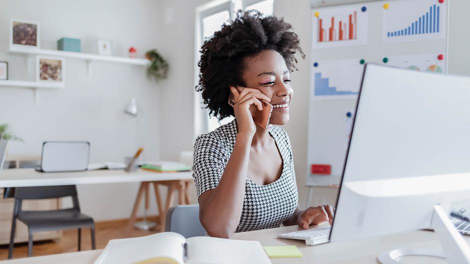 Eine Frau mit lockigem Haar spricht am Telefon und arbeitet an einem Computer in einem modernen Büro.