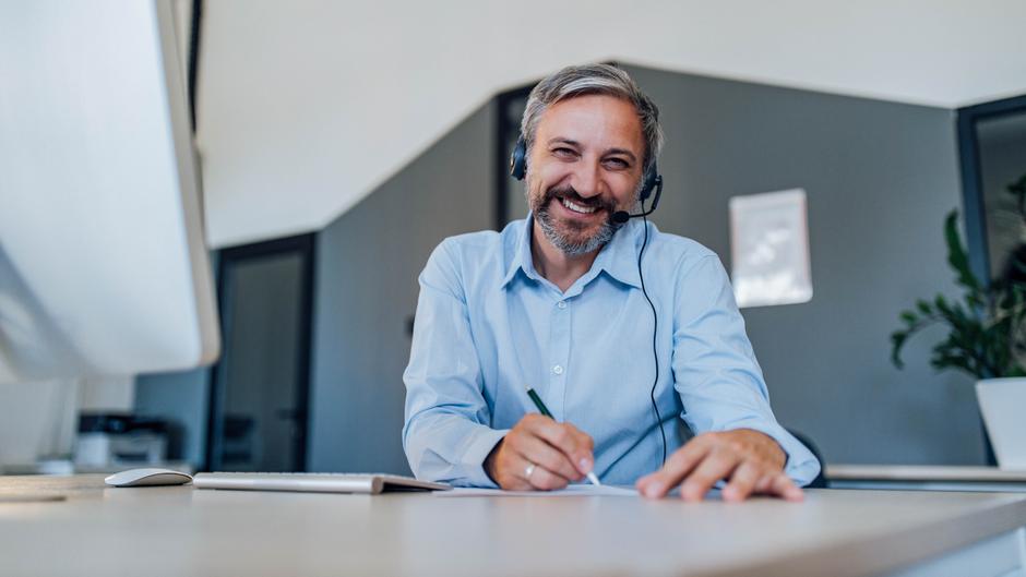 Mann mit Headset lächelt am Schreibtisch und schreibt mit einem Stift auf einem Blatt Papier.