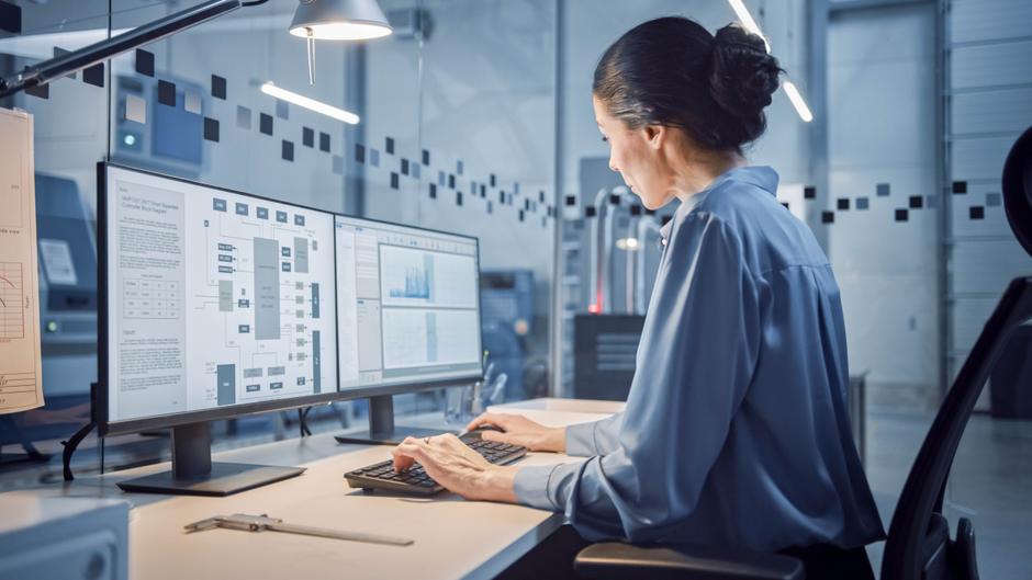 Eine Person in einem blauen Hemd arbeitet an einem Computer mit zwei Bildschirmen in einem modernen Büro.