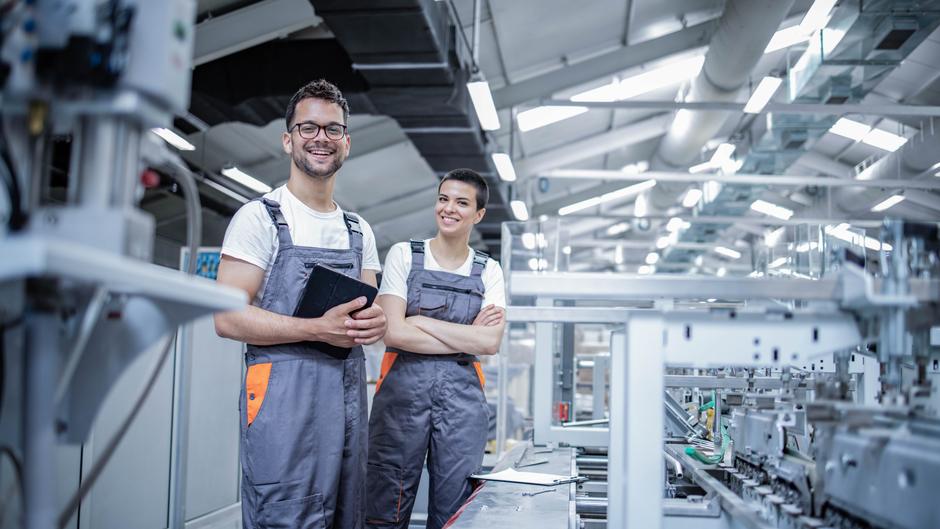 Zwei Personen in Arbeitskleidung stehen in einer modernen Produktionshalle und lächeln in die Kamera.
