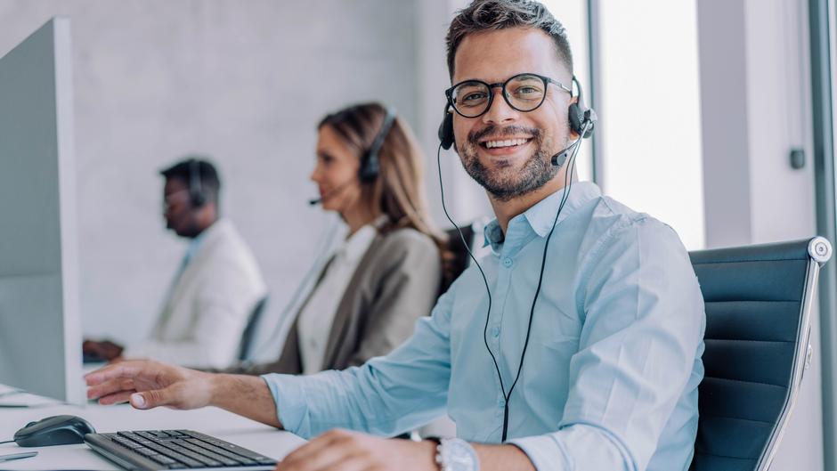 Mann mit Brille und Headset lächelt am Schreibtisch, im Hintergrund zwei weitere Personen mit Headsets.