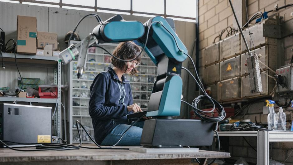 Eine Person sitzt an einem Tisch und arbeitet mit einem Roboterarm in einer Werkstatt, umgeben von Werkzeug und Materialien.