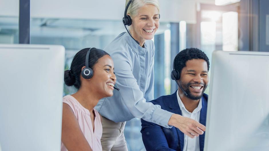 Drei Personen mit Headsets lächeln und arbeiten an Computern in einem modernen Büro.
