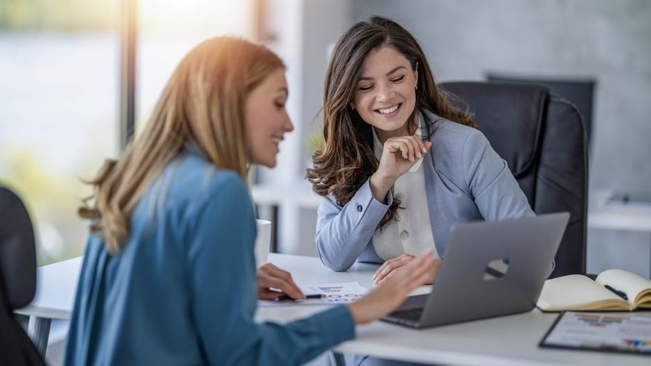 Zwei Frauen sitzen an einem Tisch und schauen lächelnd auf einen Laptop, während sie Notizen und Dokumente vor sich haben.