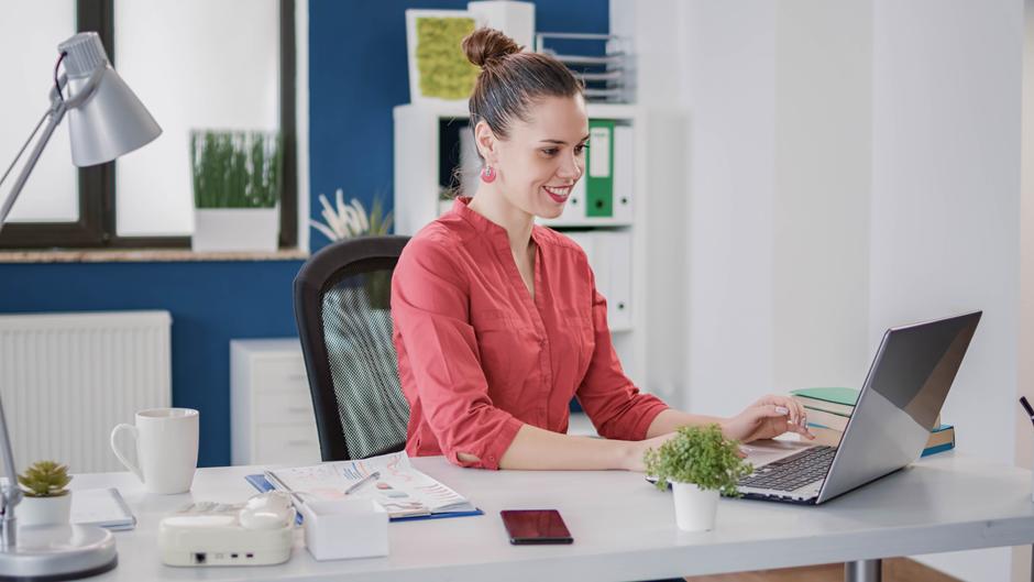 Eine Frau mit einem Dutt sitzt an einem Schreibtisch und arbeitet an einem Laptop, umgeben von Büromaterialien.