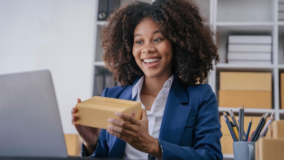 Eine lächelnde Frau mit lockigem Haar hält ein Paket in der Hand und schaut auf einen Laptop.
