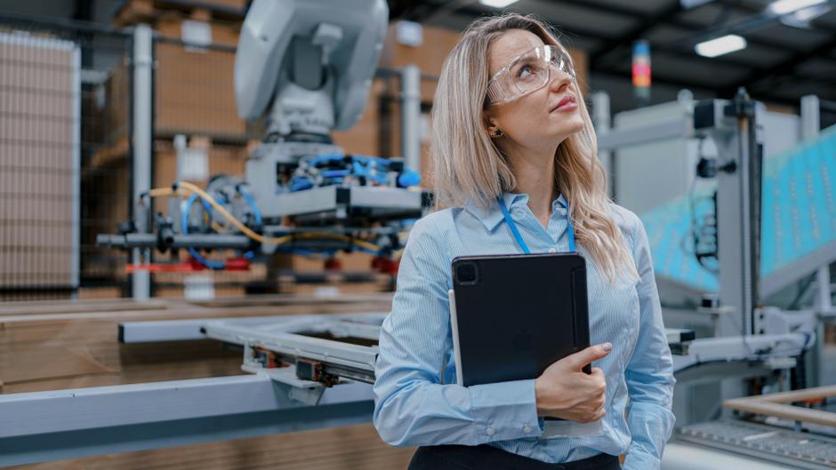 Frau mit Schutzbrille hält ein Tablet und schaut nachdenklich in einer modernen Produktionsumgebung.