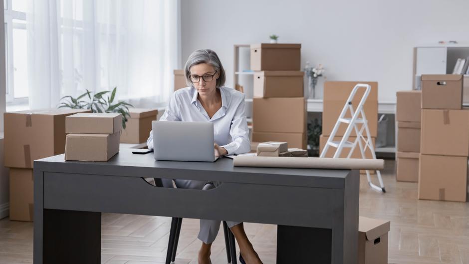 Frau mit Brille sitzt an einem Schreibtisch und arbeitet am Laptop, umgeben von Umzugskartons.